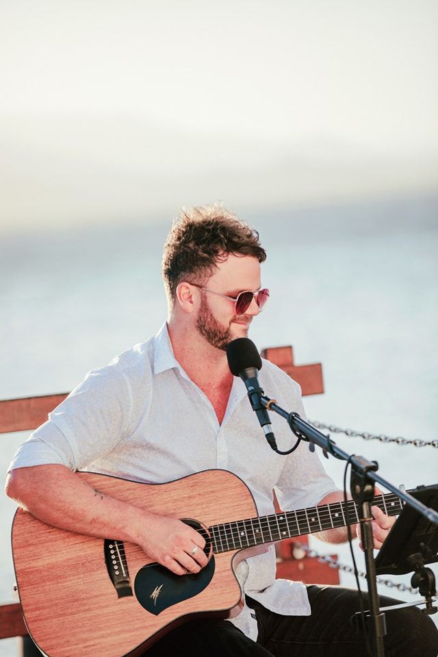 A musician playing an acoustic guitar while singing into a microphone on a seaside pier at sunset.