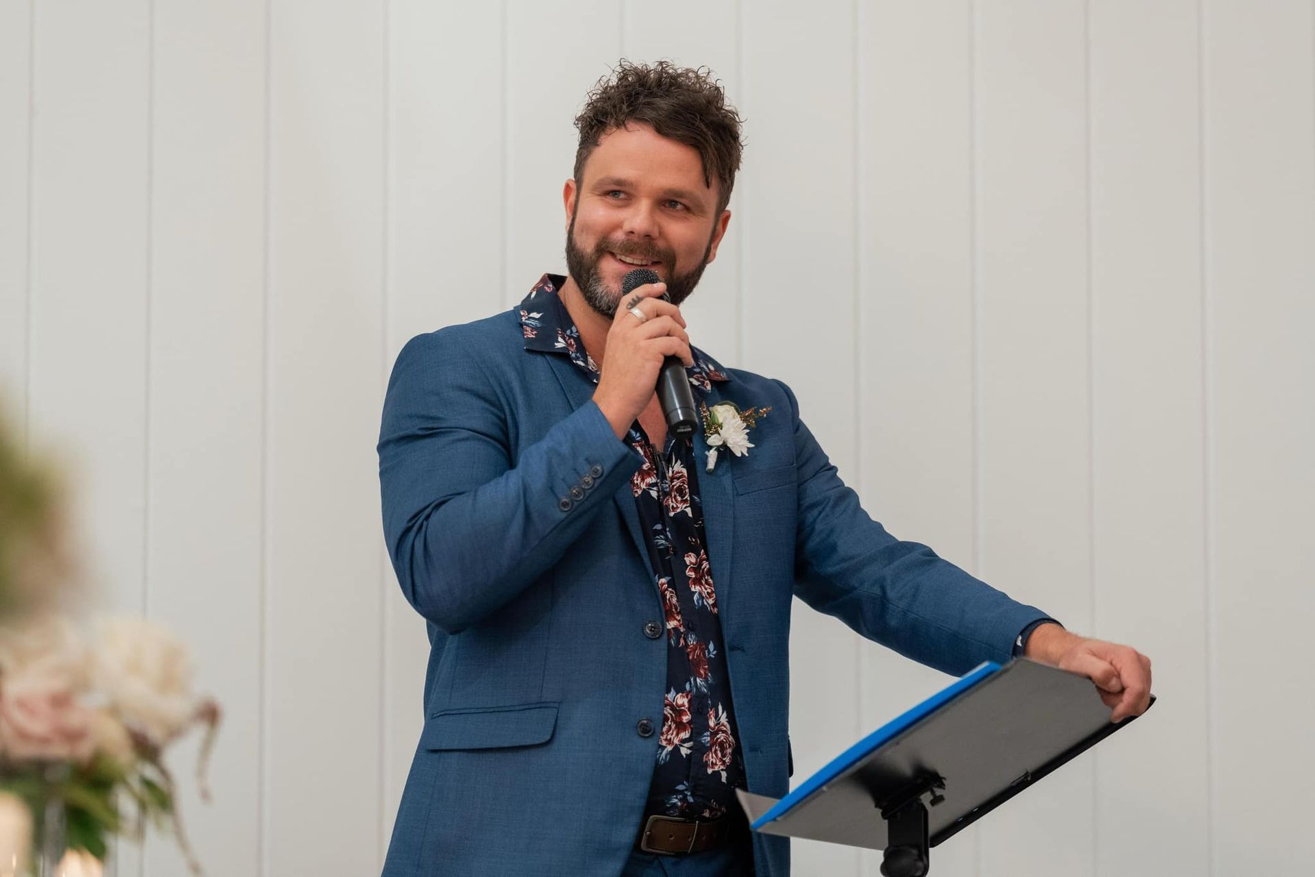 A person in a blue suit and floral shirt stands at a podium, smiling while holding a microphone to speak.