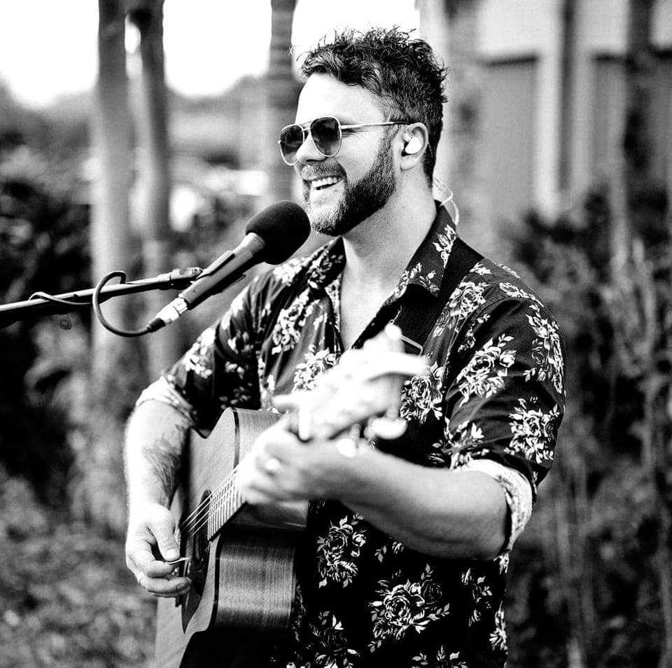 A musician wearing sunglasses and a floral shirt plays an acoustic guitar while singing into a microphone outdoors.