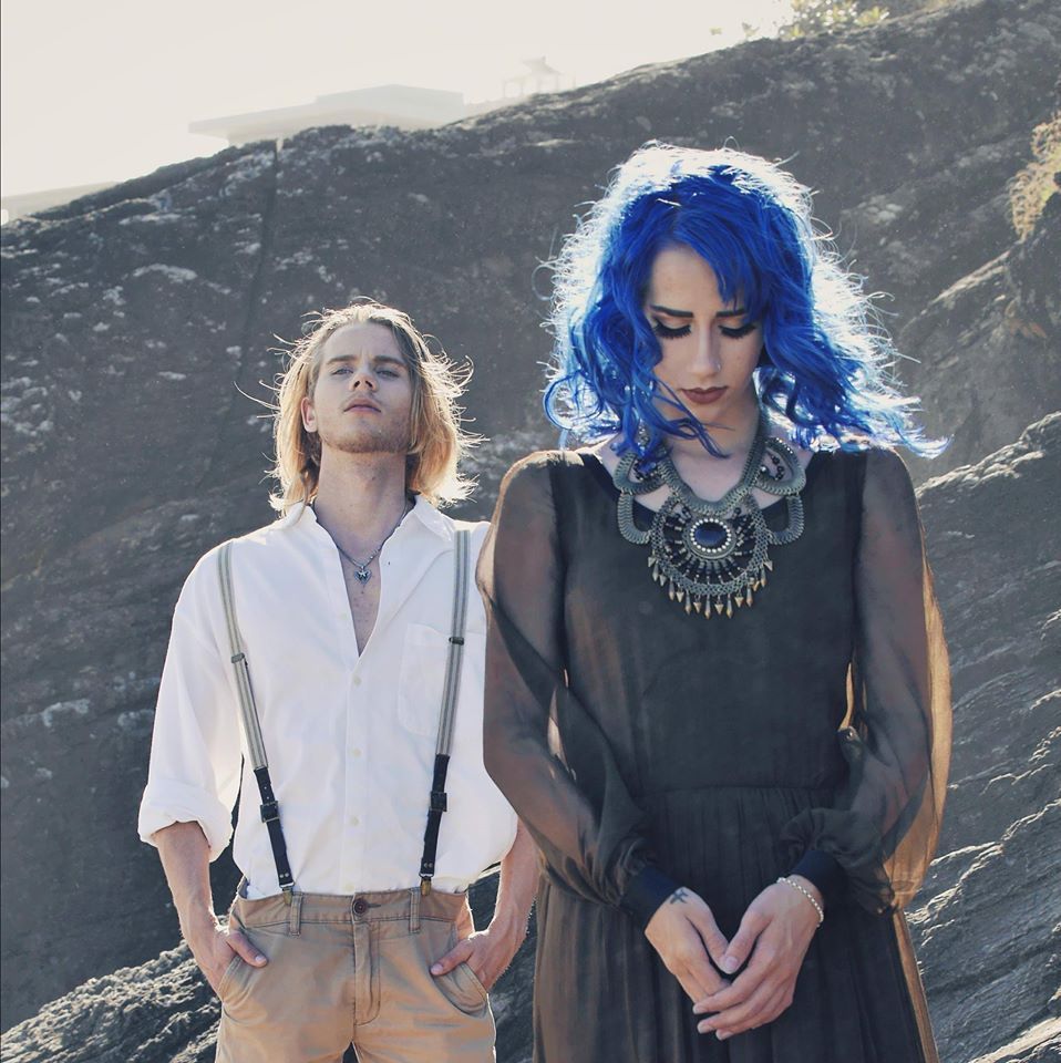 A person with bright blue hair and a dark dress stands before another in a white shirt and suspenders on a rocky hill.