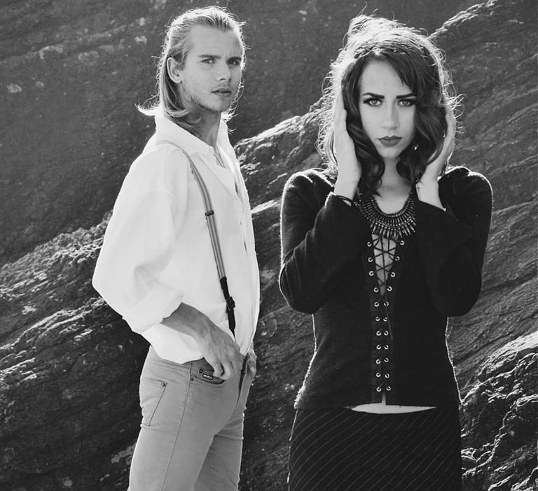 Black-and-white portrait of two people standing against a rocky backdrop, posing with serious expressions.