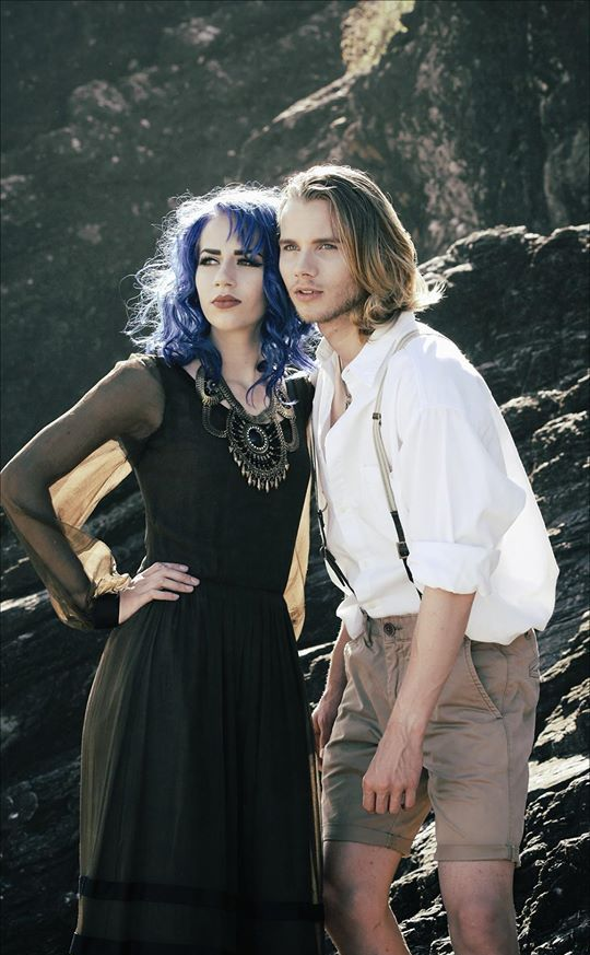 A person with vibrant blue hair and a black gown stands beside a person in a white shirt and khaki shorts on rocky ground.