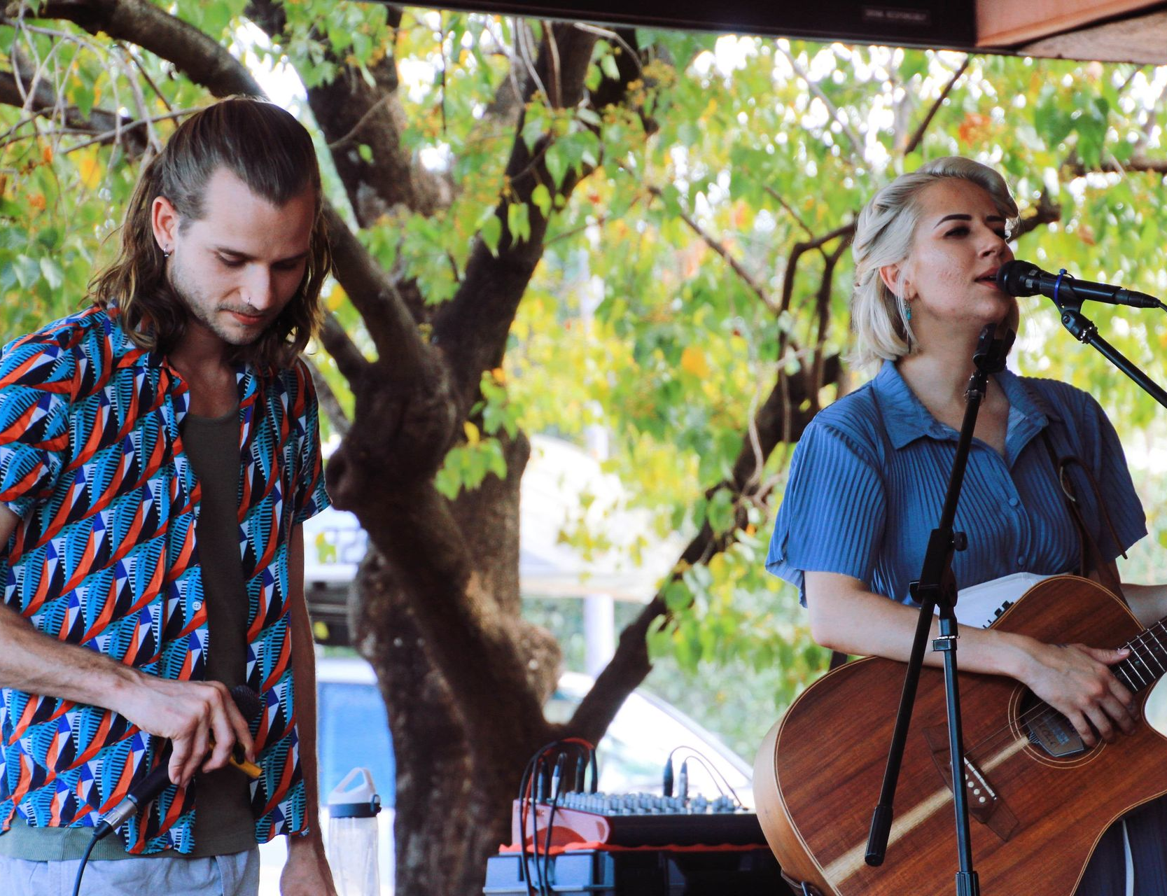Two musicians performing outdoors, one playing acoustic guitar at a microphone and the other holding a small instrument.