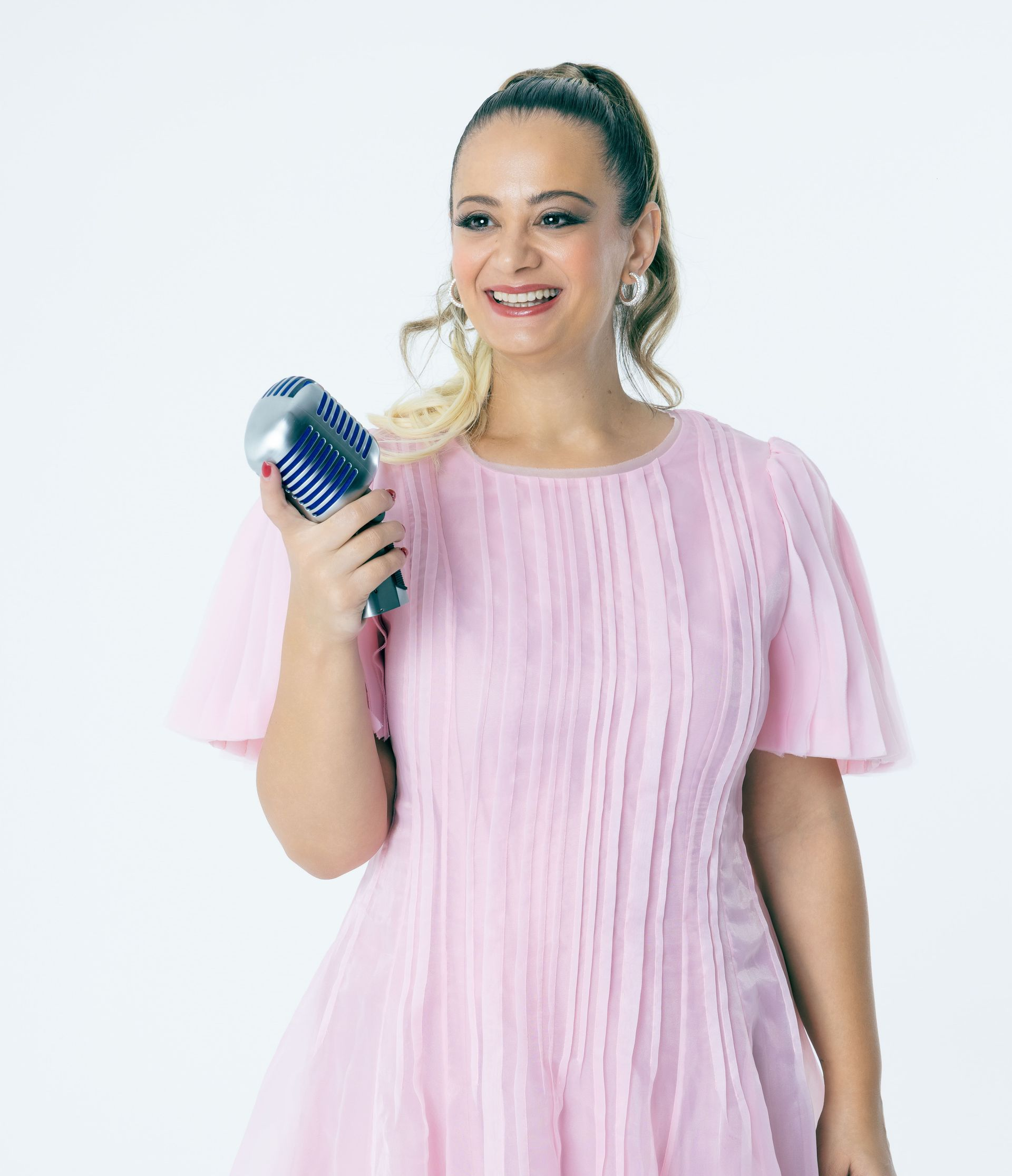 Woman in pink dress holds a microphone, smiling against a white background.