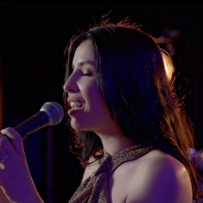 A person singing into a handheld microphone in a dimly lit setting with blue and warm lighting on their face.