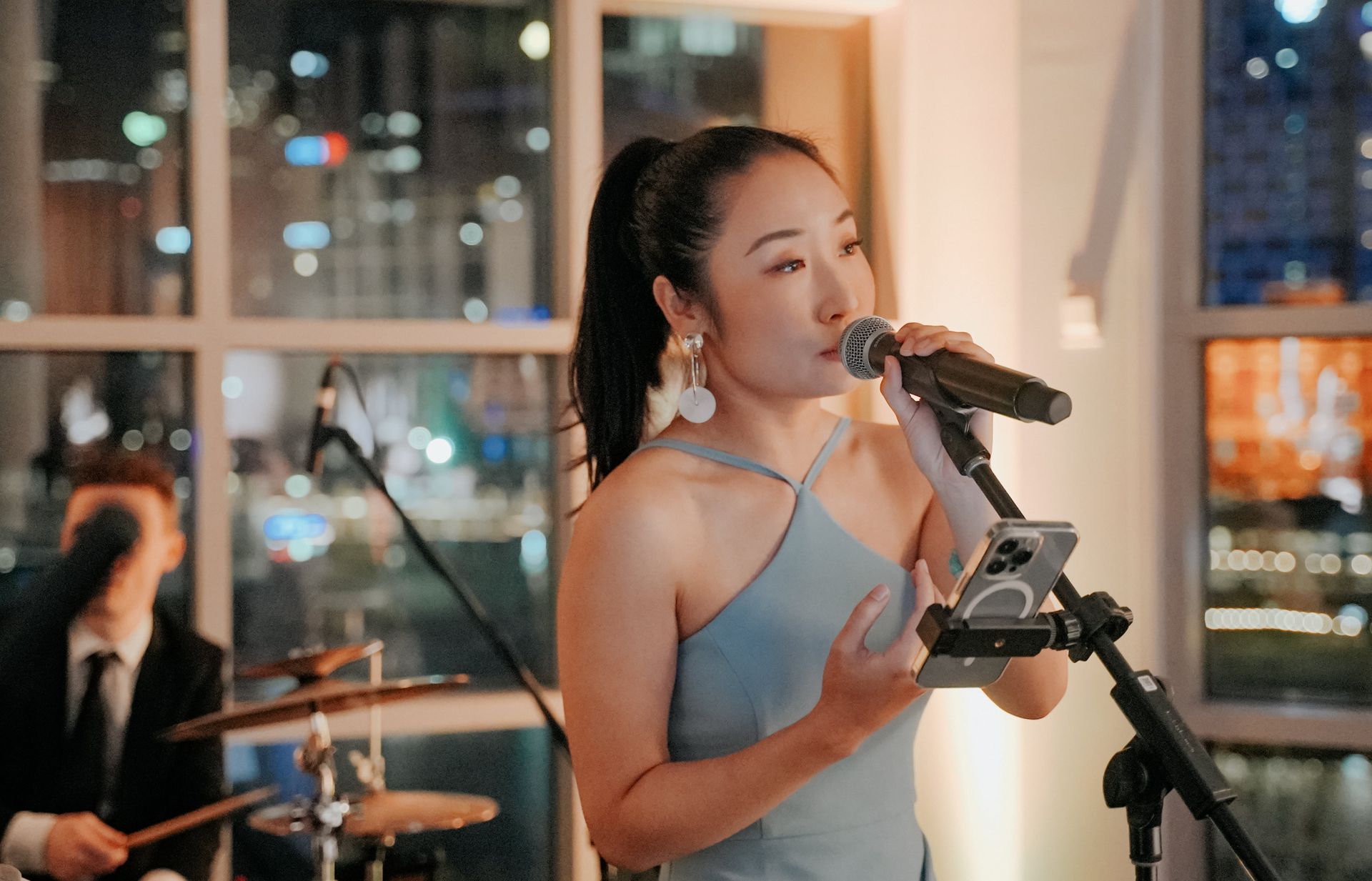 Woman singing into a microphone on stage, with a drummer in the background, near a large window at night.