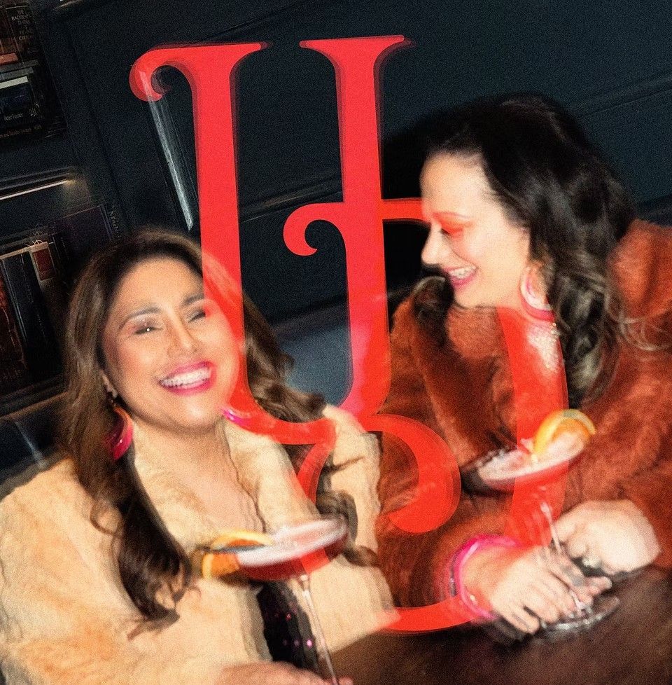 Two people in coats holding cocktails, smiling at a bar