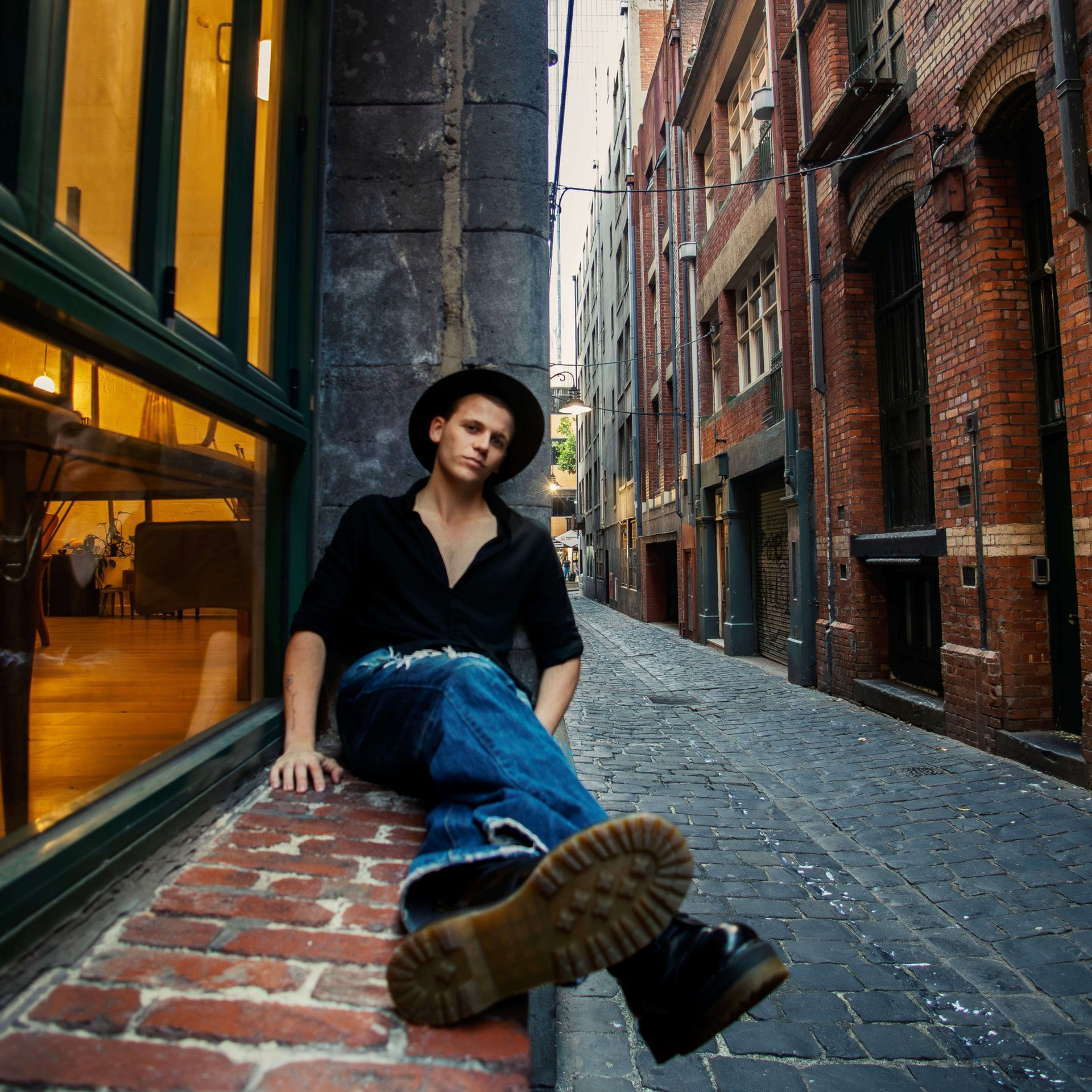Person in a black hat and shirt sits on a brick ledge in a narrow cobblestone alley.