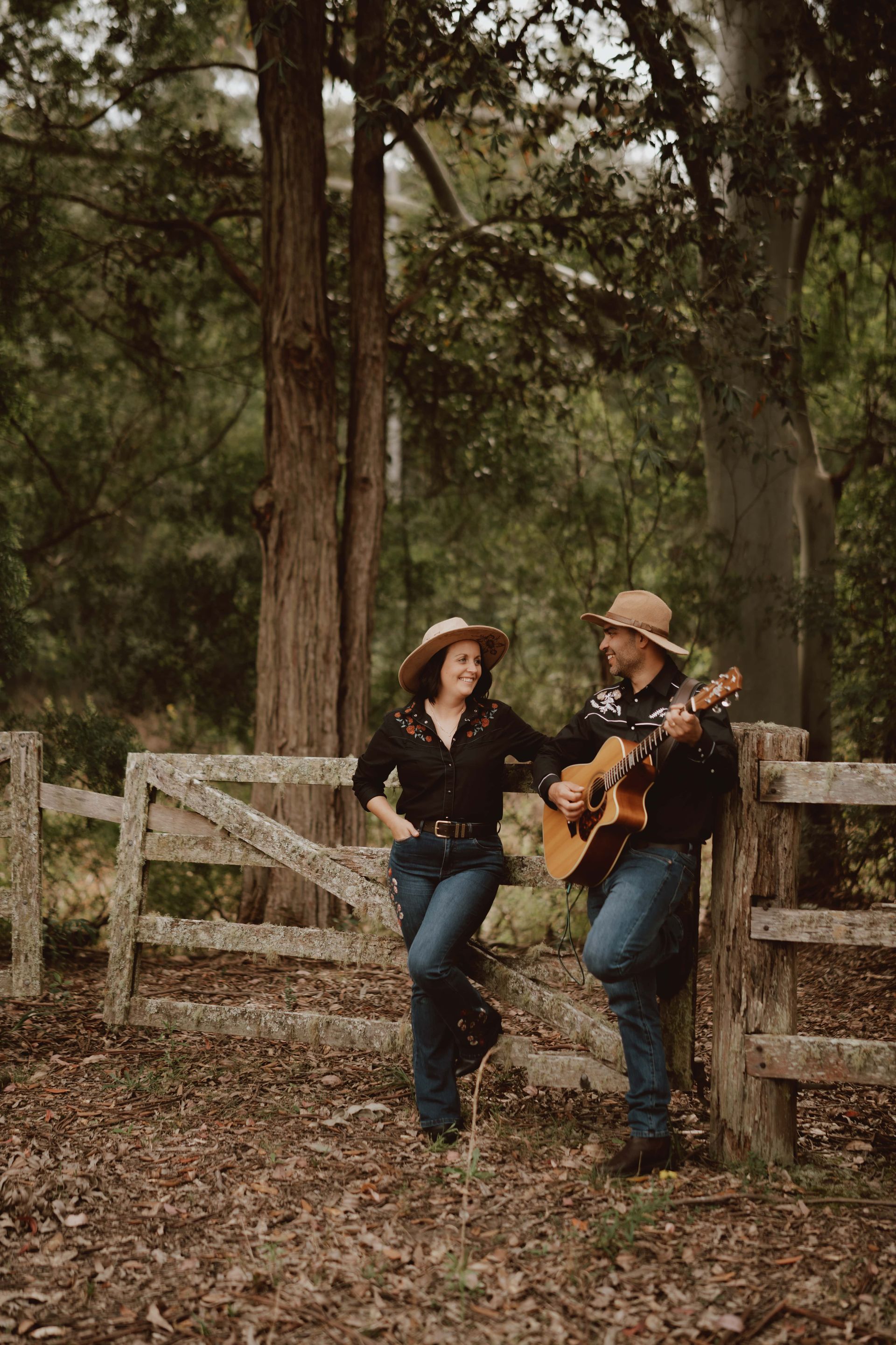 Two people in western attire lean on a rustic wooden fence in a forest, one playing an acoustic guitar.