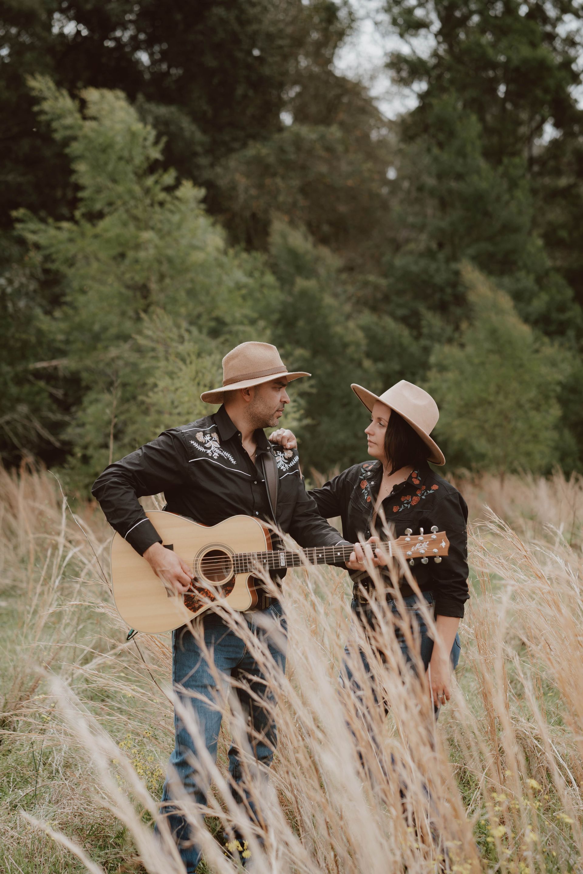 Two people in cowboy hats and matching black shirts stand in a field of tall grass, one playing an acoustic guitar.