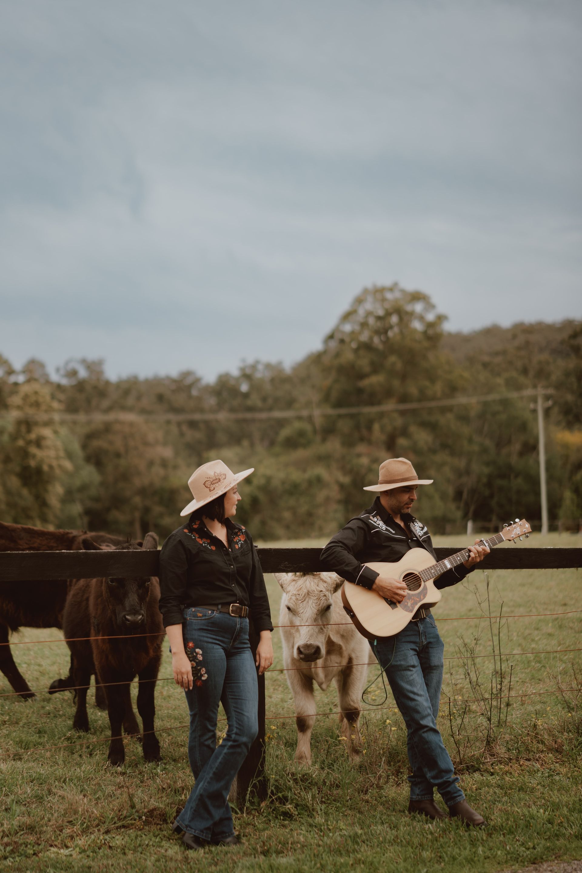 Two people wearing hats and western-style clothing stand in a field with cows, one playing an acoustic guitar.
