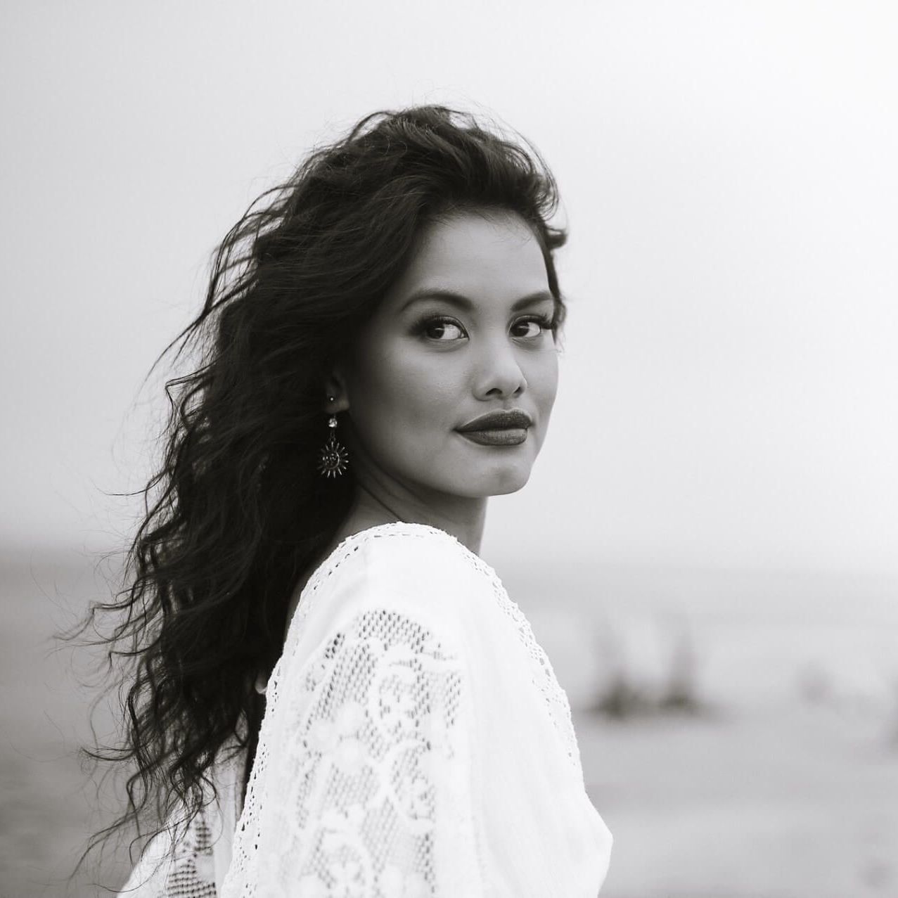 A person with long, dark, wavy hair, wearing a white lace garment, looks back over their shoulder against a misty backdrop.