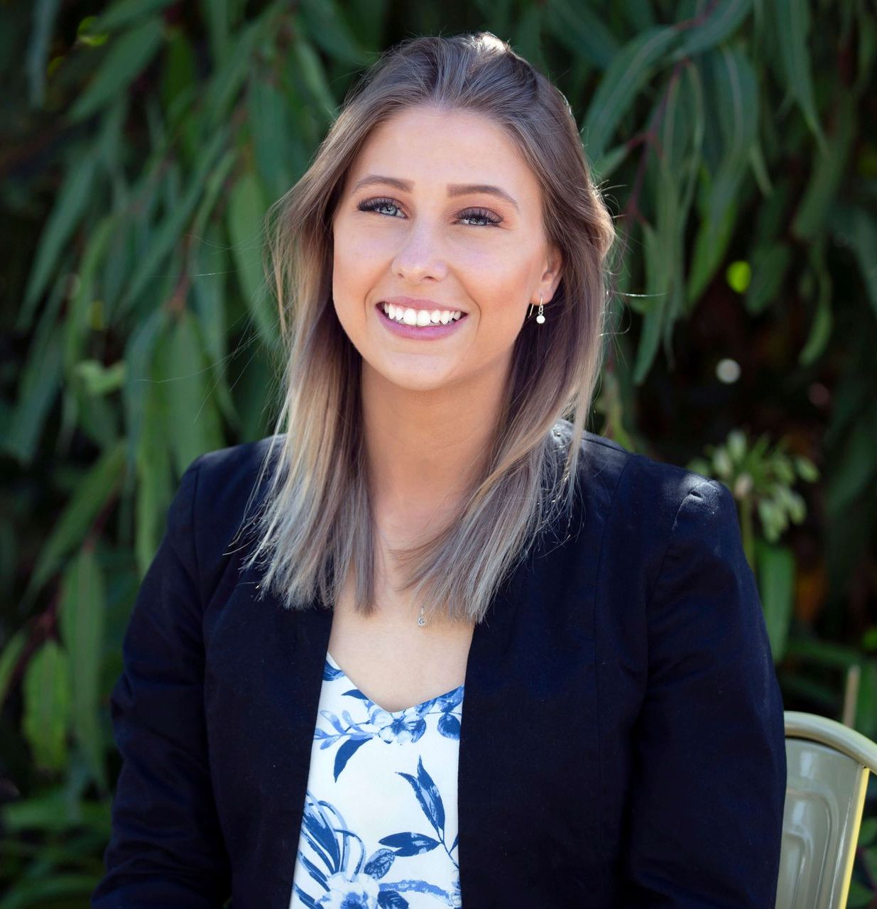 Woman with blonde hair, smiling, wearing a black blazer over a blue and white floral top. Green background.