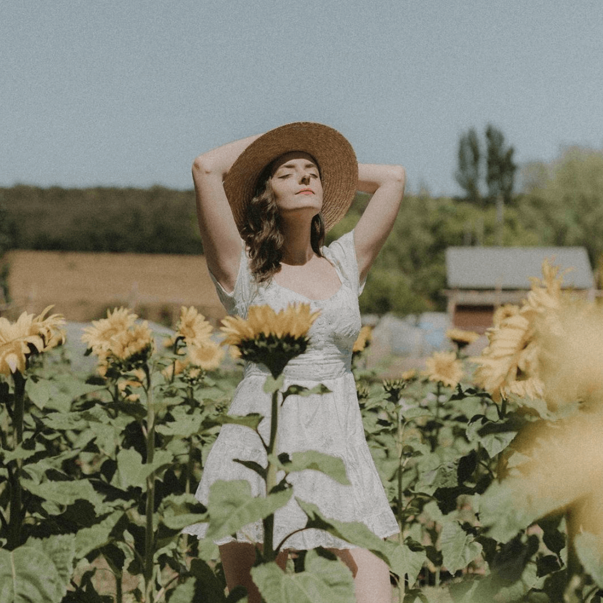 A woman in a white dress and straw hat is standing in a field of sunflowers.