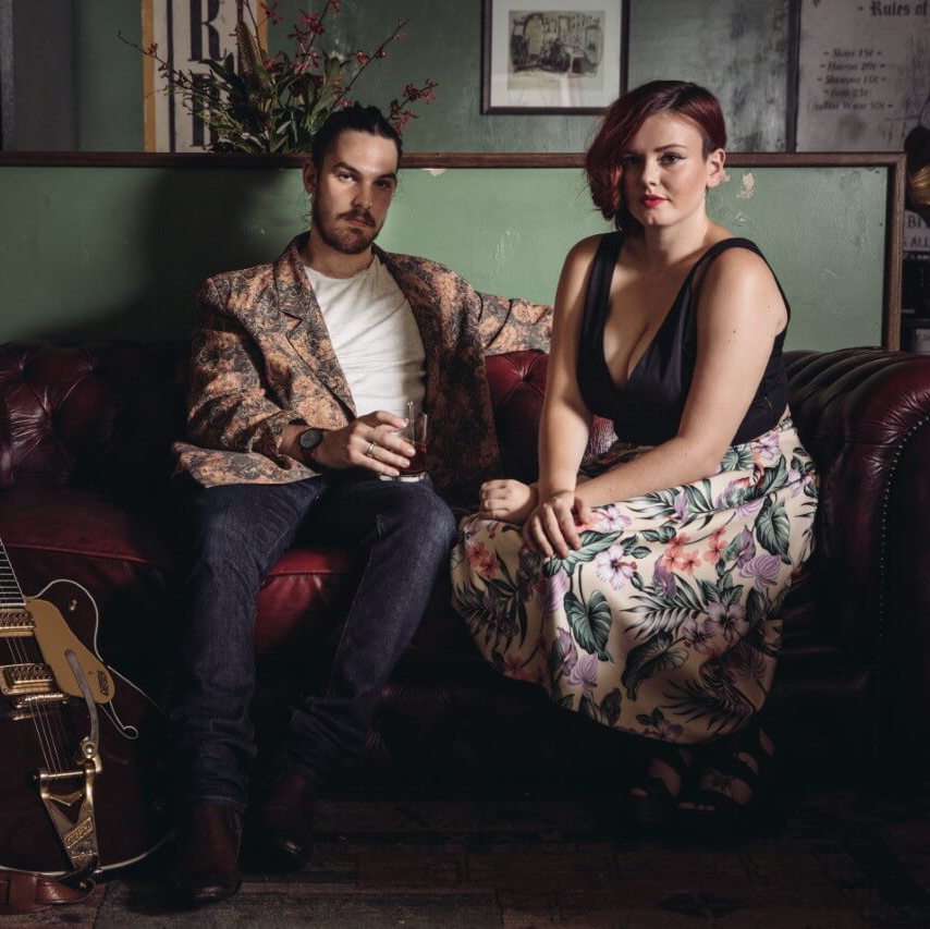 A man and woman sit on a red leather sofa in front of a green wall, with an acoustic guitar resting on the floor.