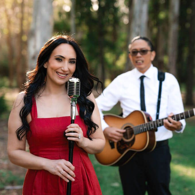 Woman in a red dress sings into a vintage microphone while a guitarist plays behind her outdoors.
