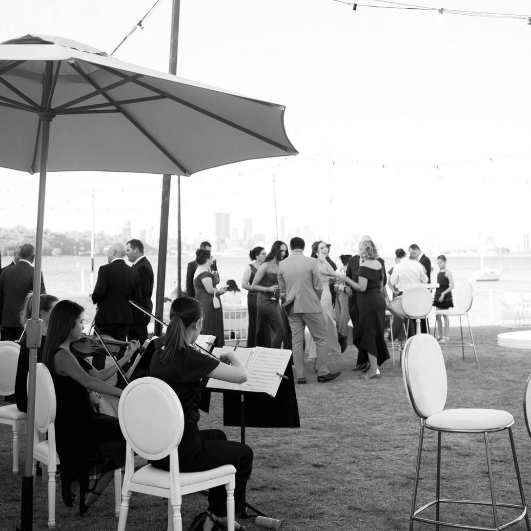 Two musicians play violins under an umbrella at an outdoor event with guests socializing by the water in the background.