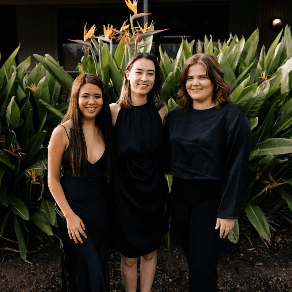 Three individuals stand side-by-side wearing black outfits in front of a lush green plant with orange flowers.
