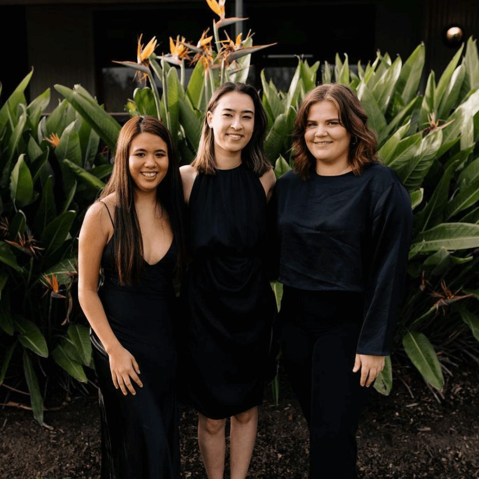 Three people in black outfits stand smiling together in front of green, leafy plants.