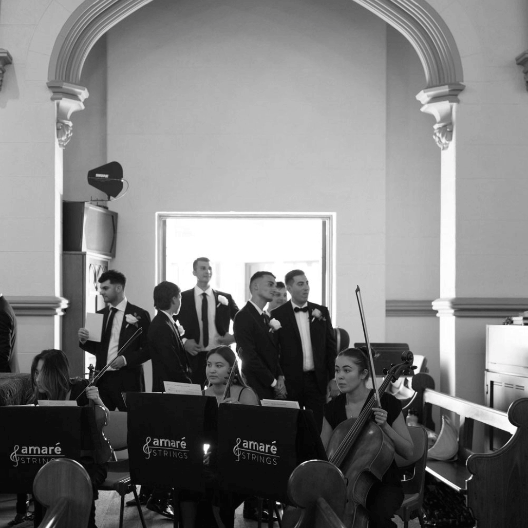 Musicians in black outfits perform in a church archway as formally dressed guests stand in the background.
