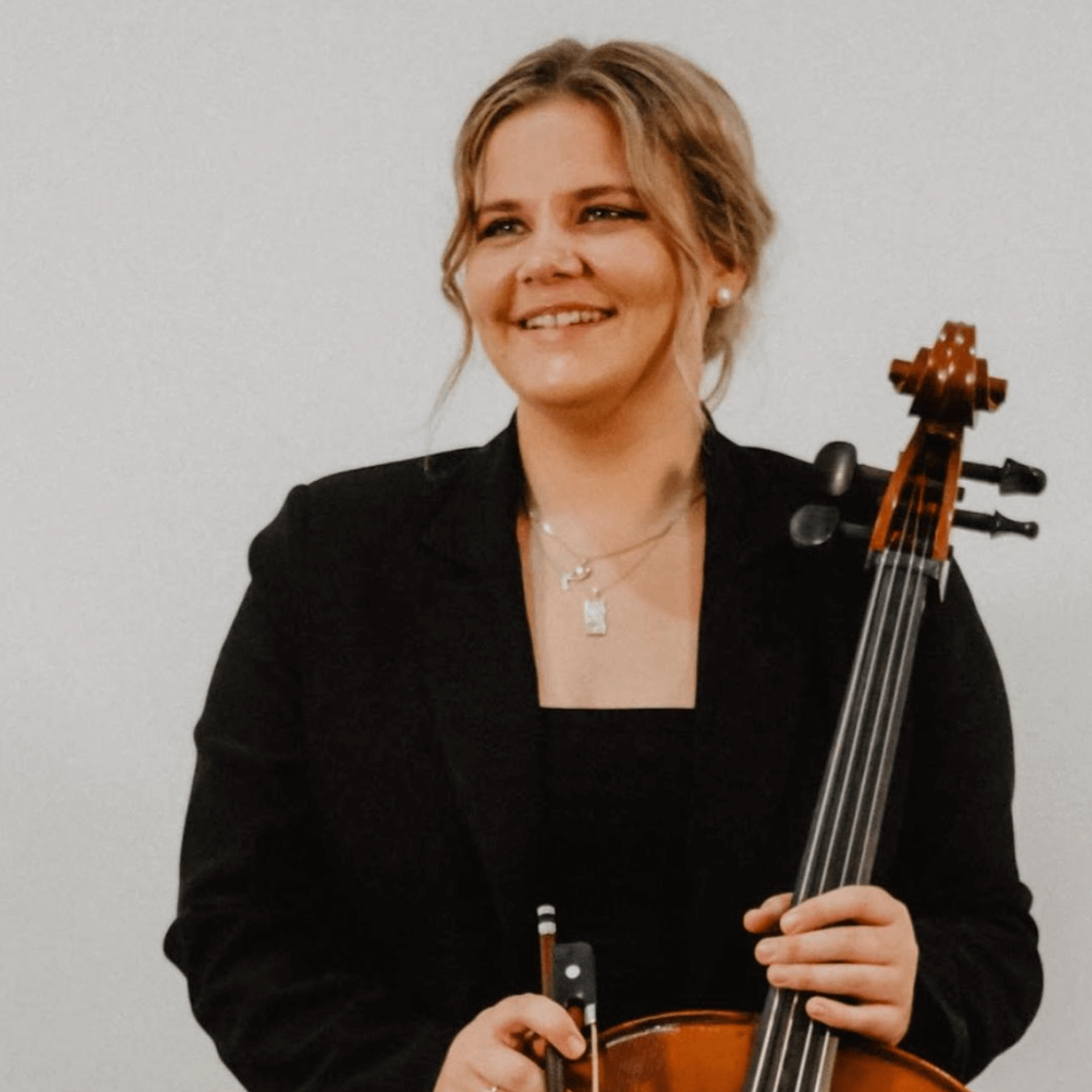 A smiling musician in a black blazer stands holding a cello and a bow against a plain white background.