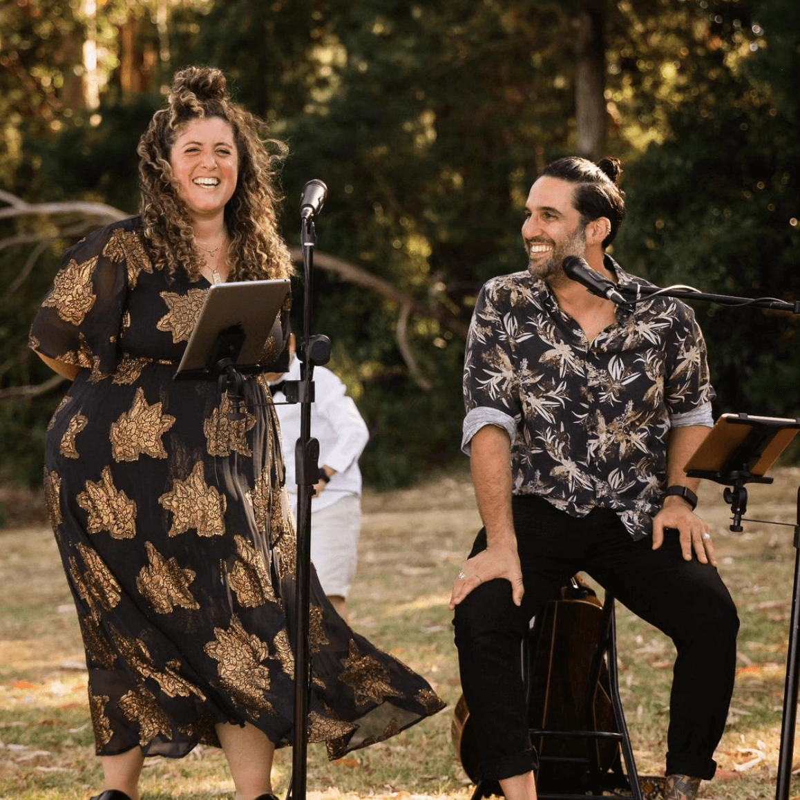 Two people performing outdoors with microphones, smiling. One has a tablet, the other an acoustic guitar.