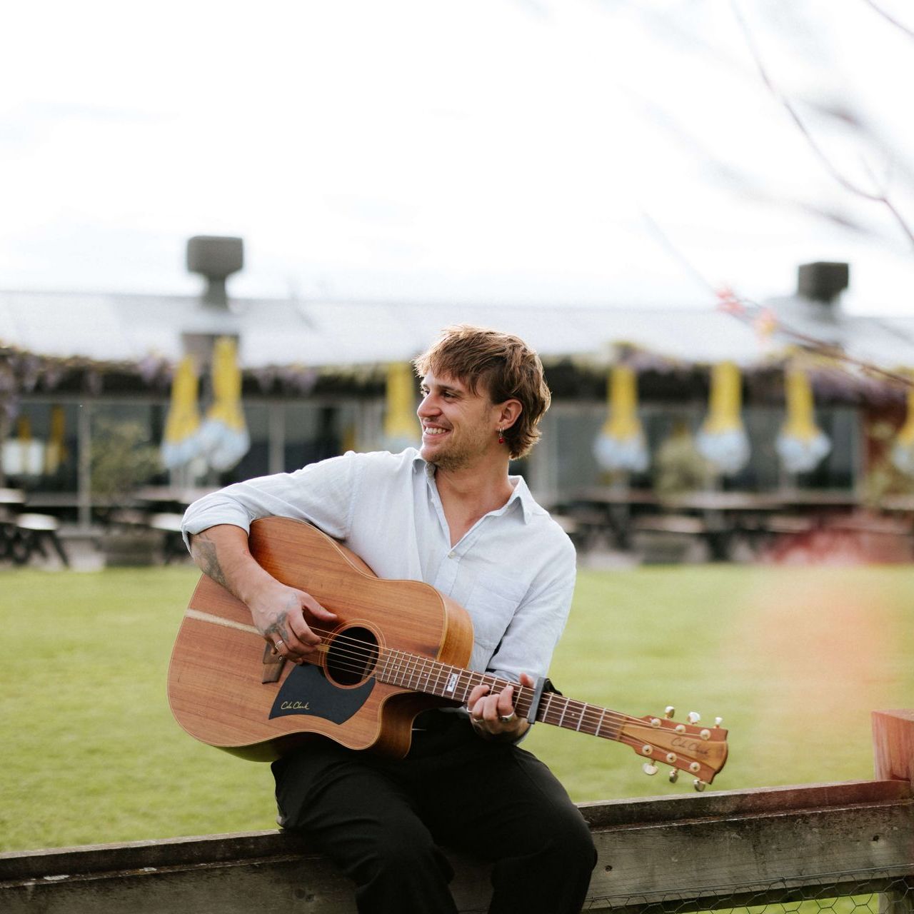 Man sitting on a fence, playing an acoustic guitar. He smiles, outdoors in front of a building.