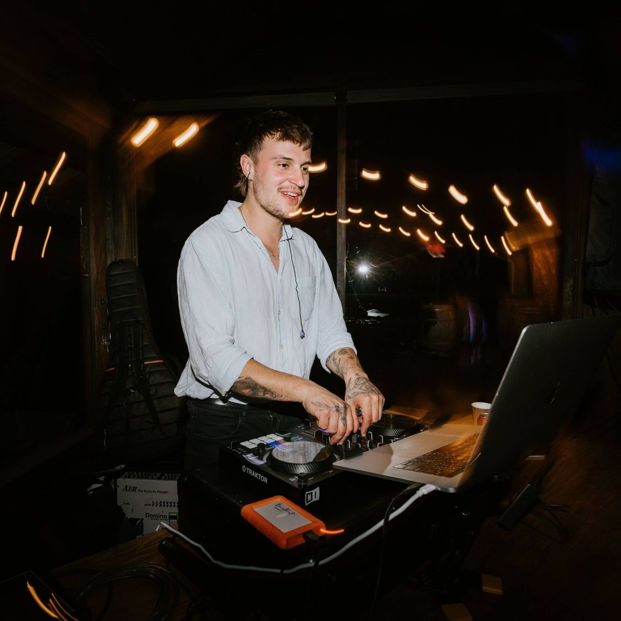 DJ smiling and mixing music at a dark venue. He wears a light shirt, has tattoos, and stands in front of a laptop.