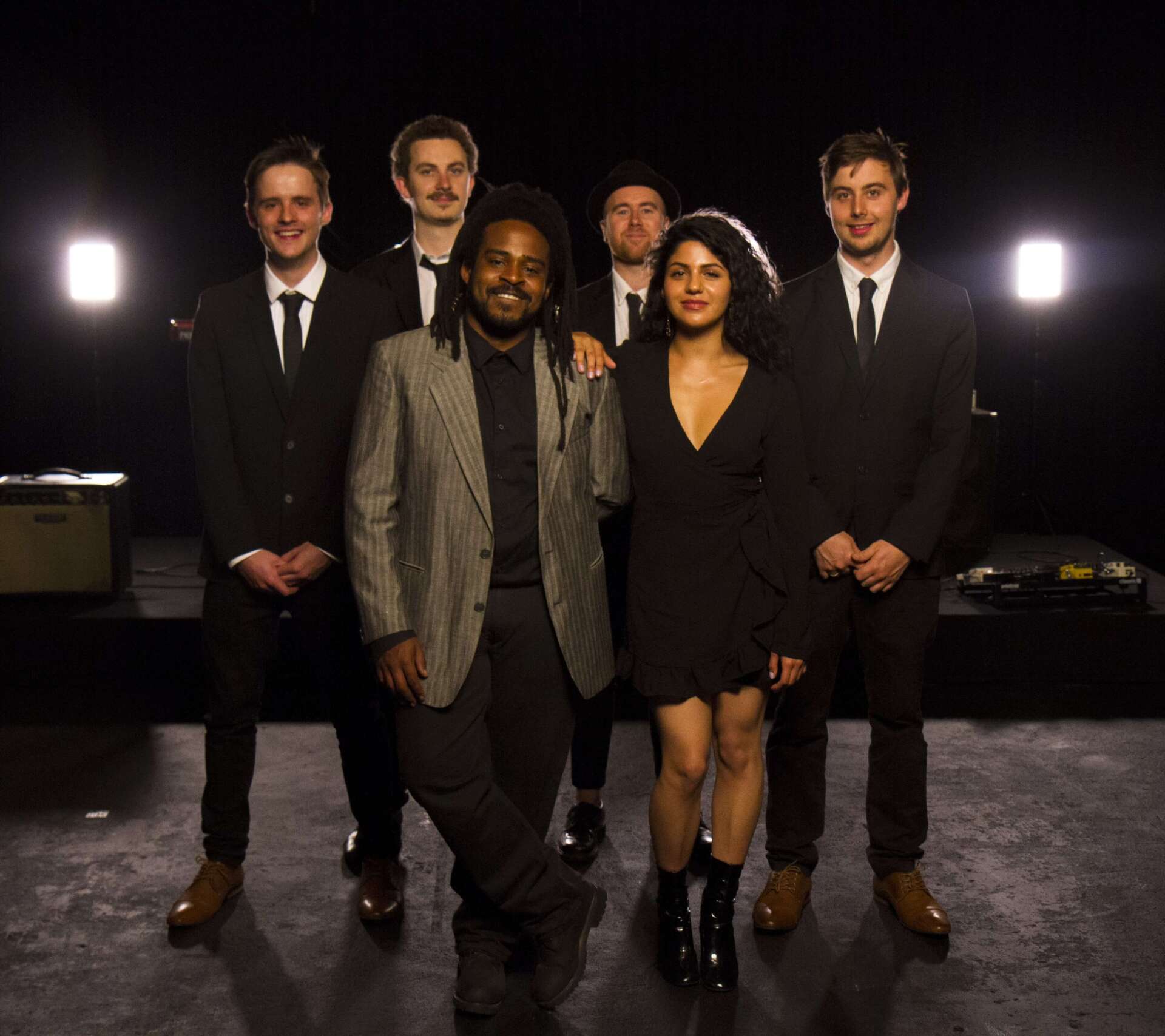 Band of seven in suits poses on a stage. One in a gray jacket leans forward, others stand. Black background.