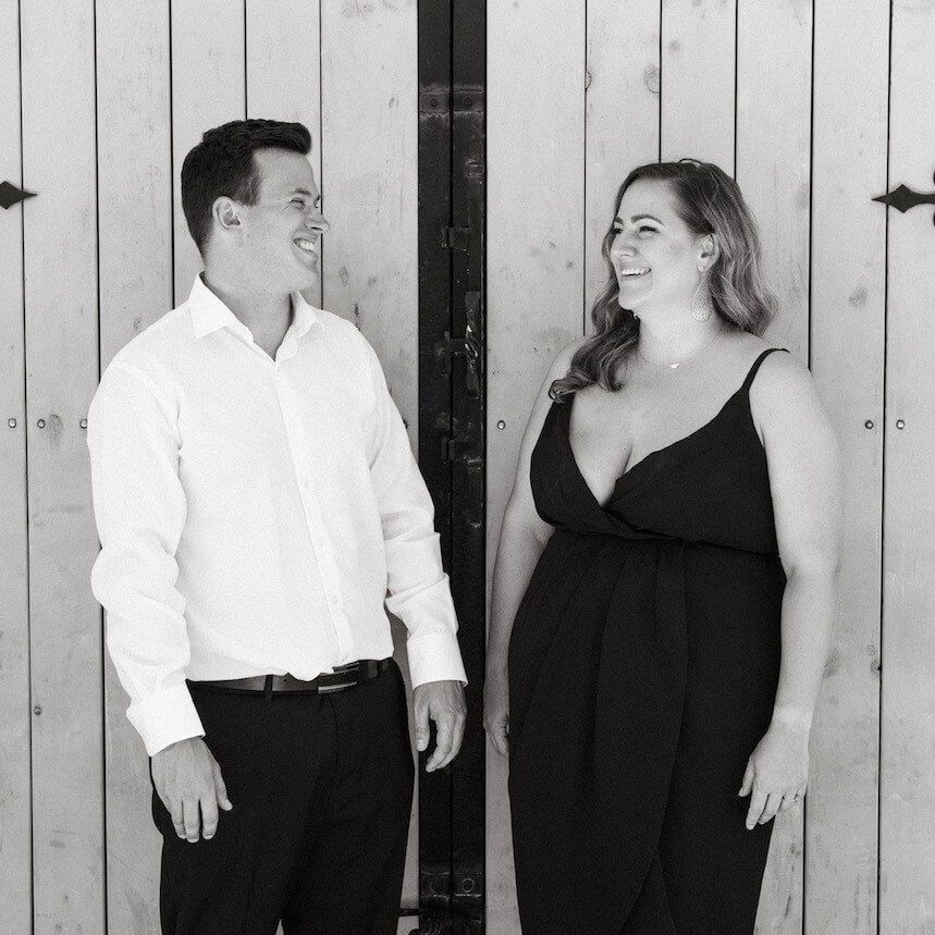 Couple smiling, standing by a wooden door. The woman wears a black dress, the man a white shirt and black pants.