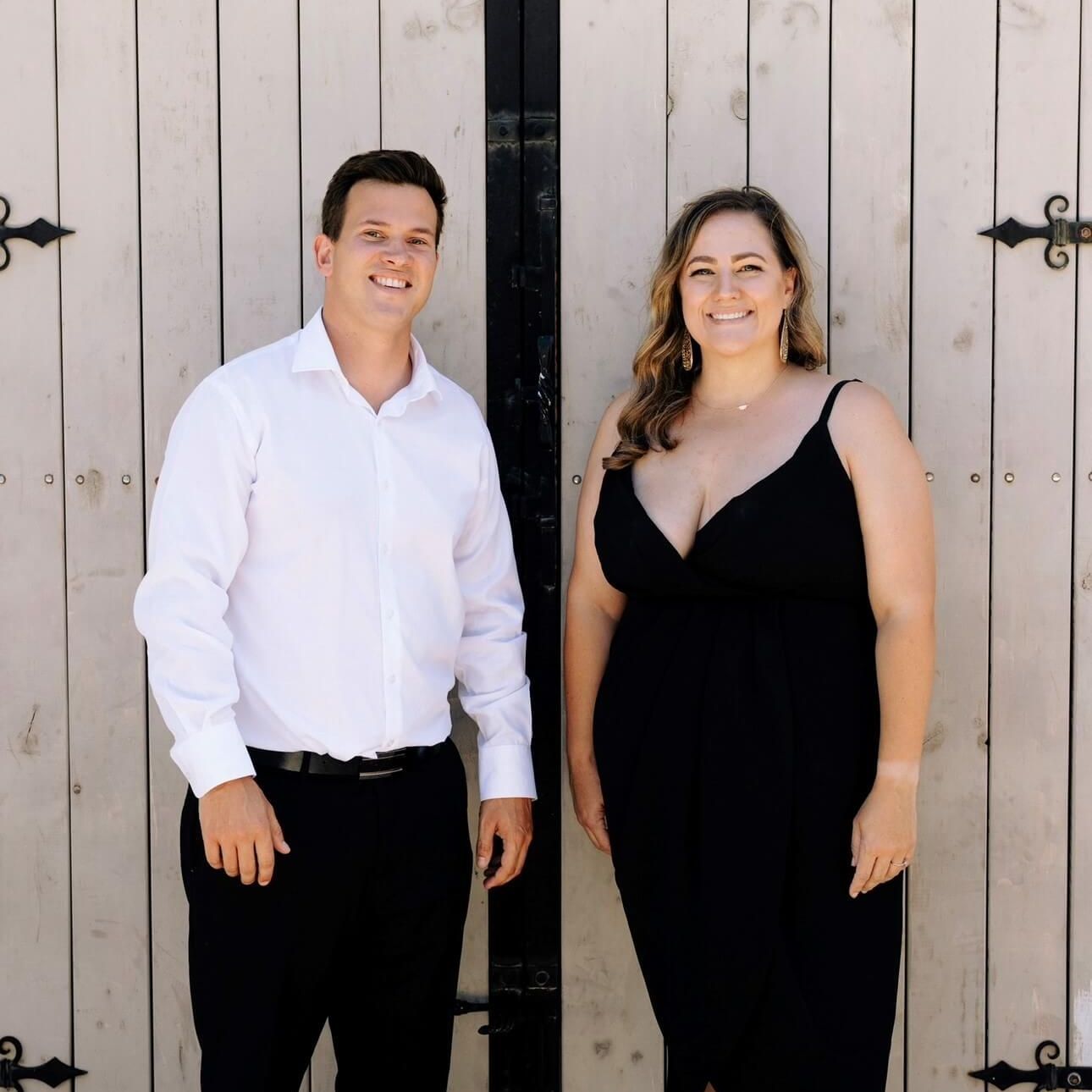 Man and woman standing in front of a white wooden door. Man in a white shirt and black pants, woman in a black dress.