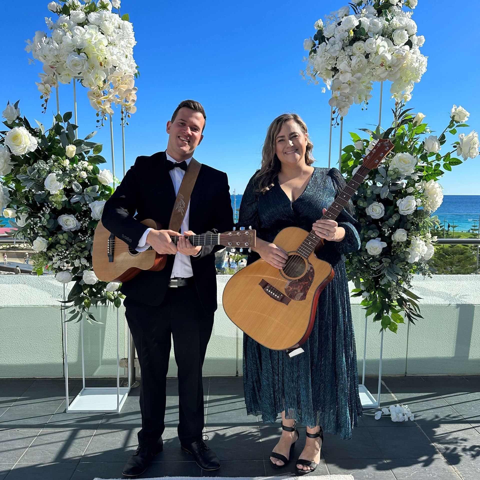 Two people with guitars perform outdoors near floral arrangements; ocean in the background.
