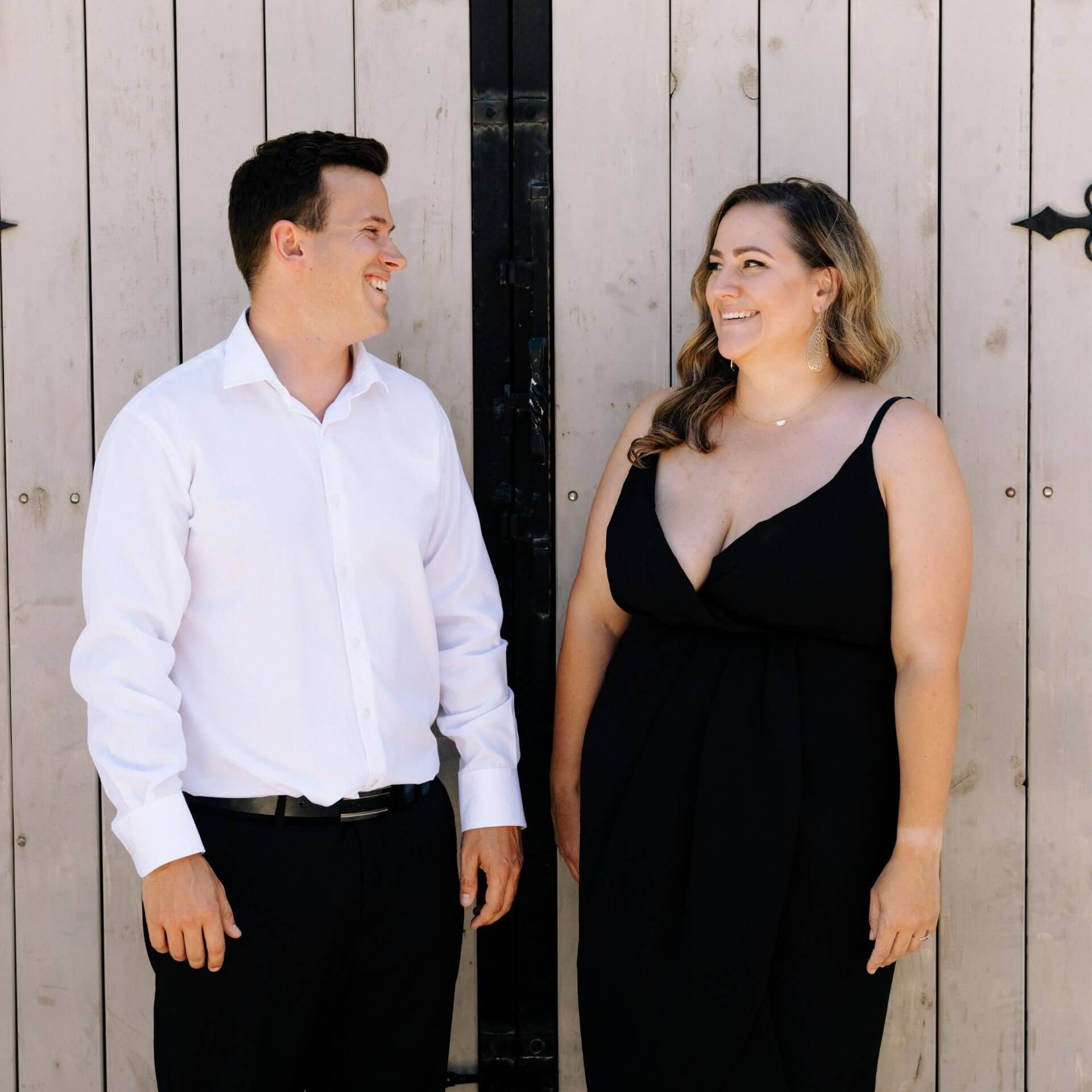 Man and woman smiling, standing in front of a weathered, light wooden door. The woman wears a black dress. The man wears a white shirt.