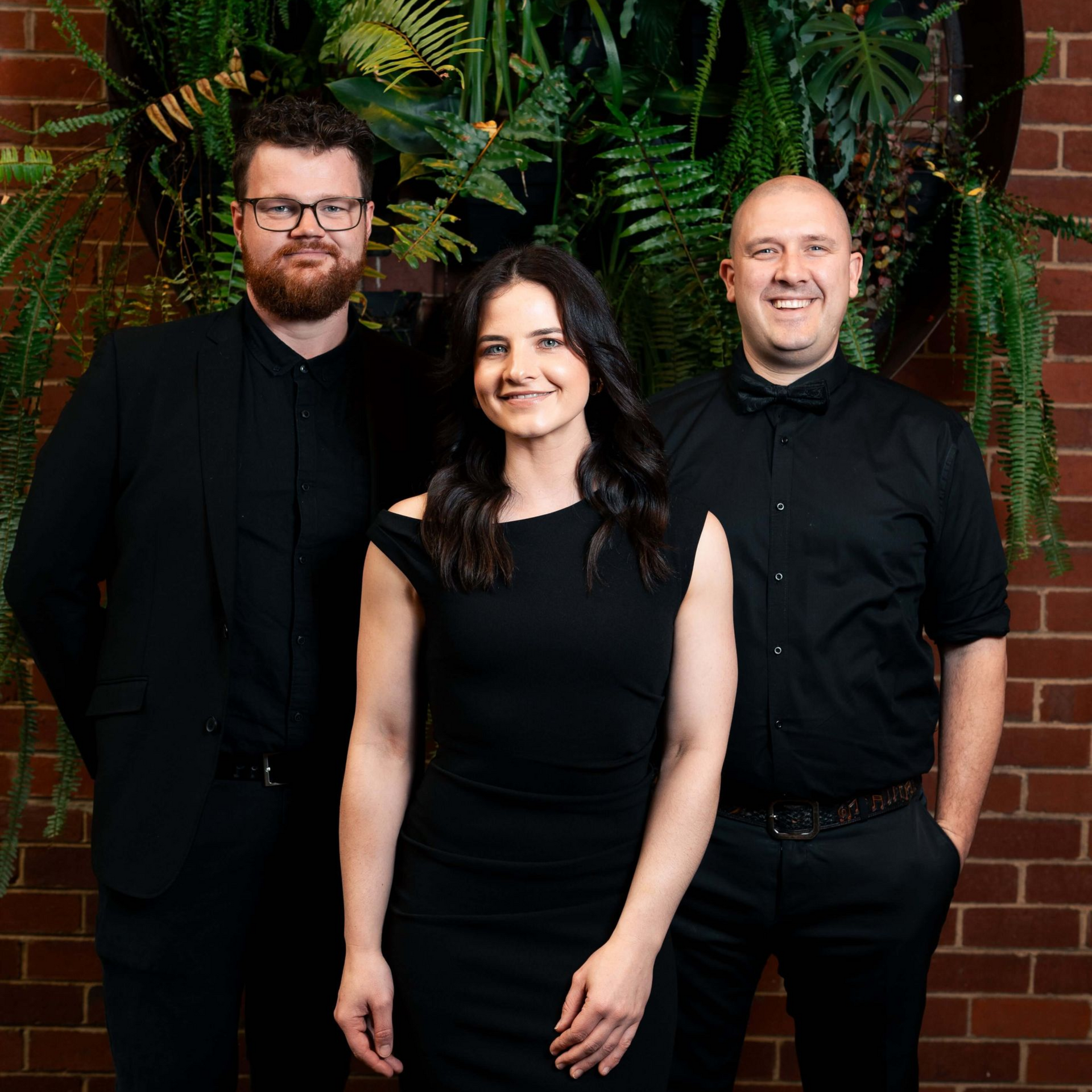 Three people in black attire pose in front of a brick wall with greenery.
