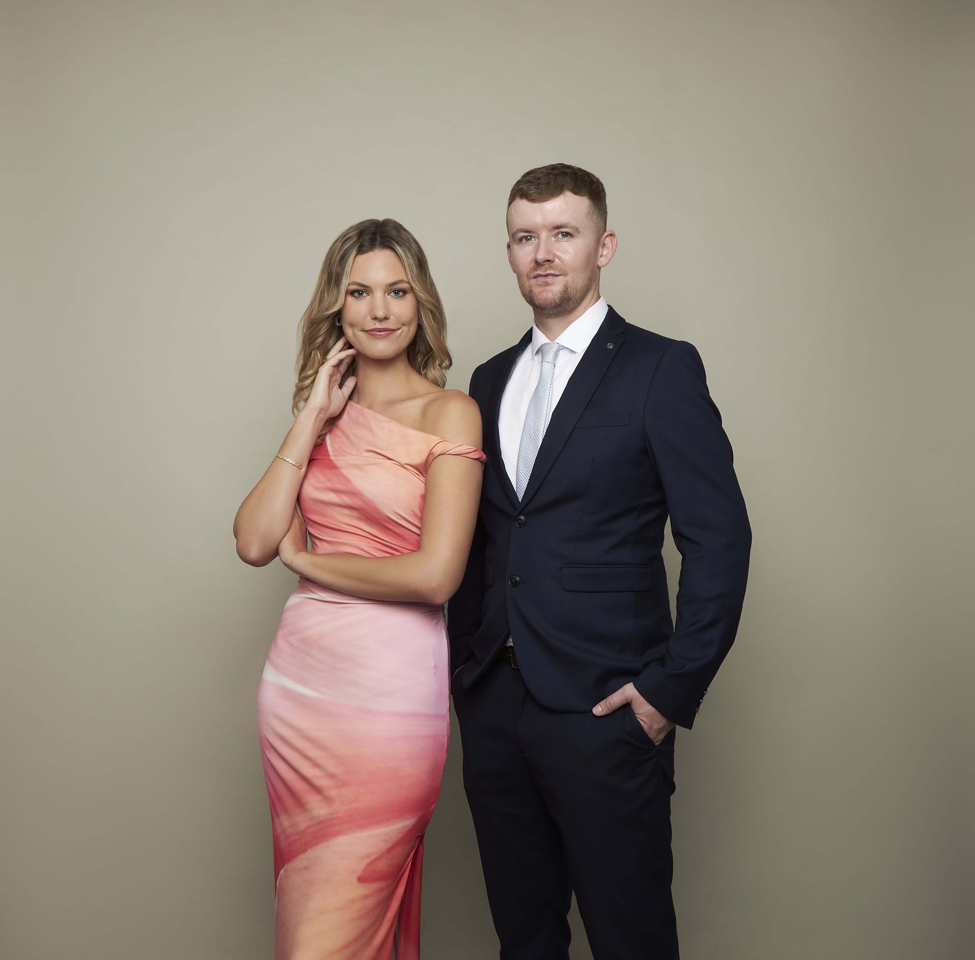 A man in a suit standing next to a person in a pink and orange gradient evening dress against a plain neutral background.
