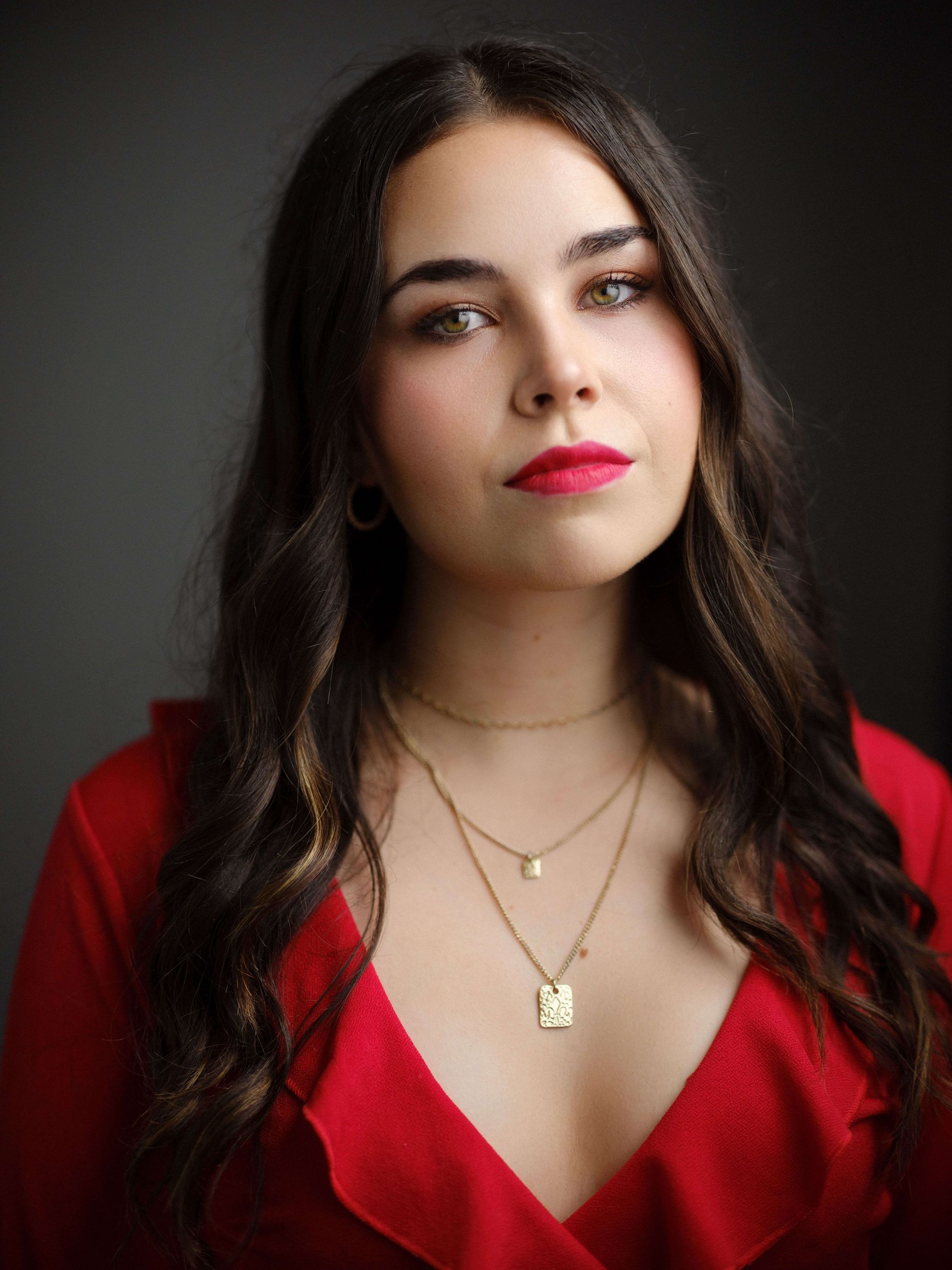 Woman with long, wavy brown hair, wearing a red top and gold necklaces, looking towards the camera.