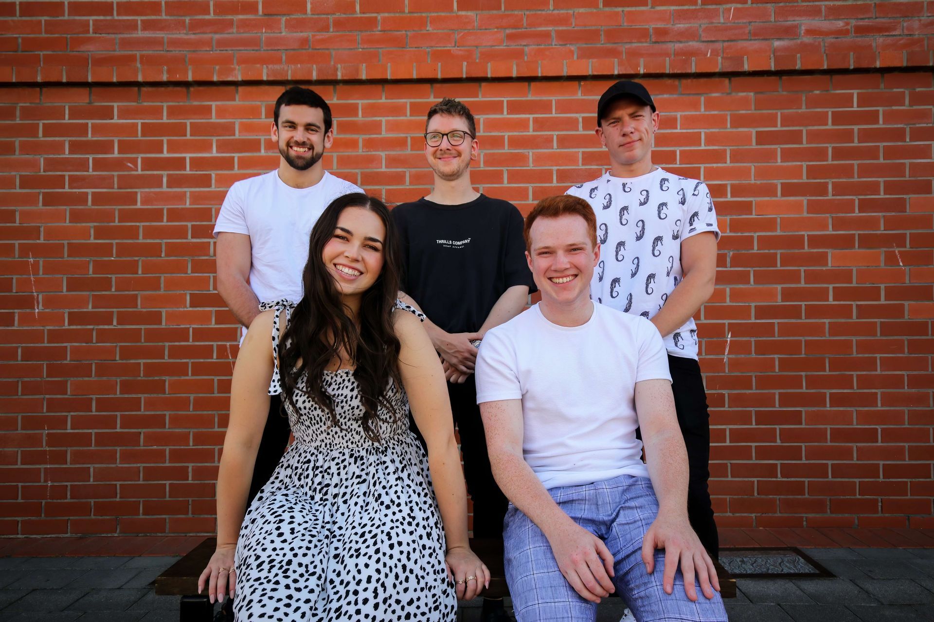 Five people smiling, posing in front of a brick wall. Two seated in front row, three standing behind.