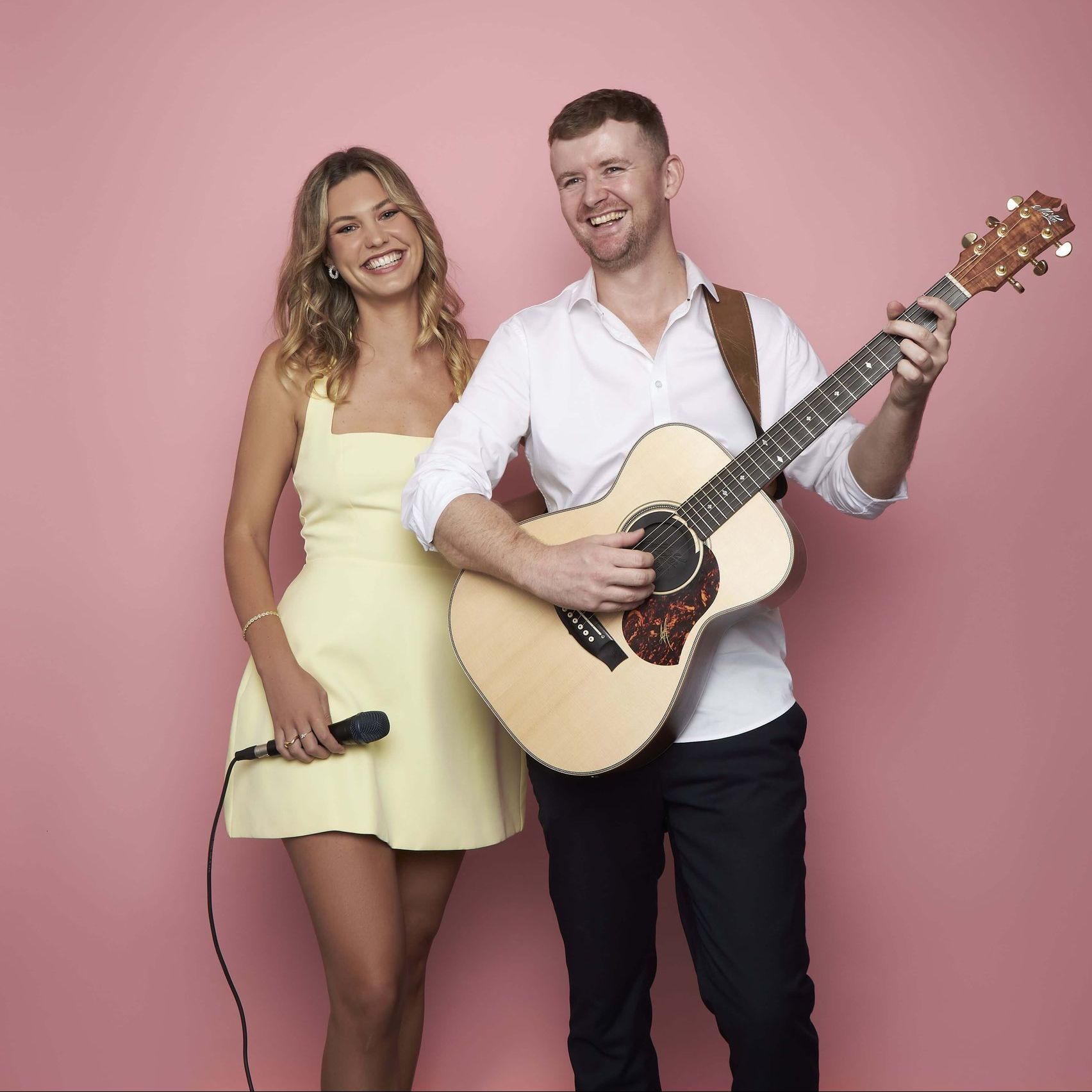 A smiling woman holding a microphone stands next to a man playing an acoustic guitar against a solid pink background.