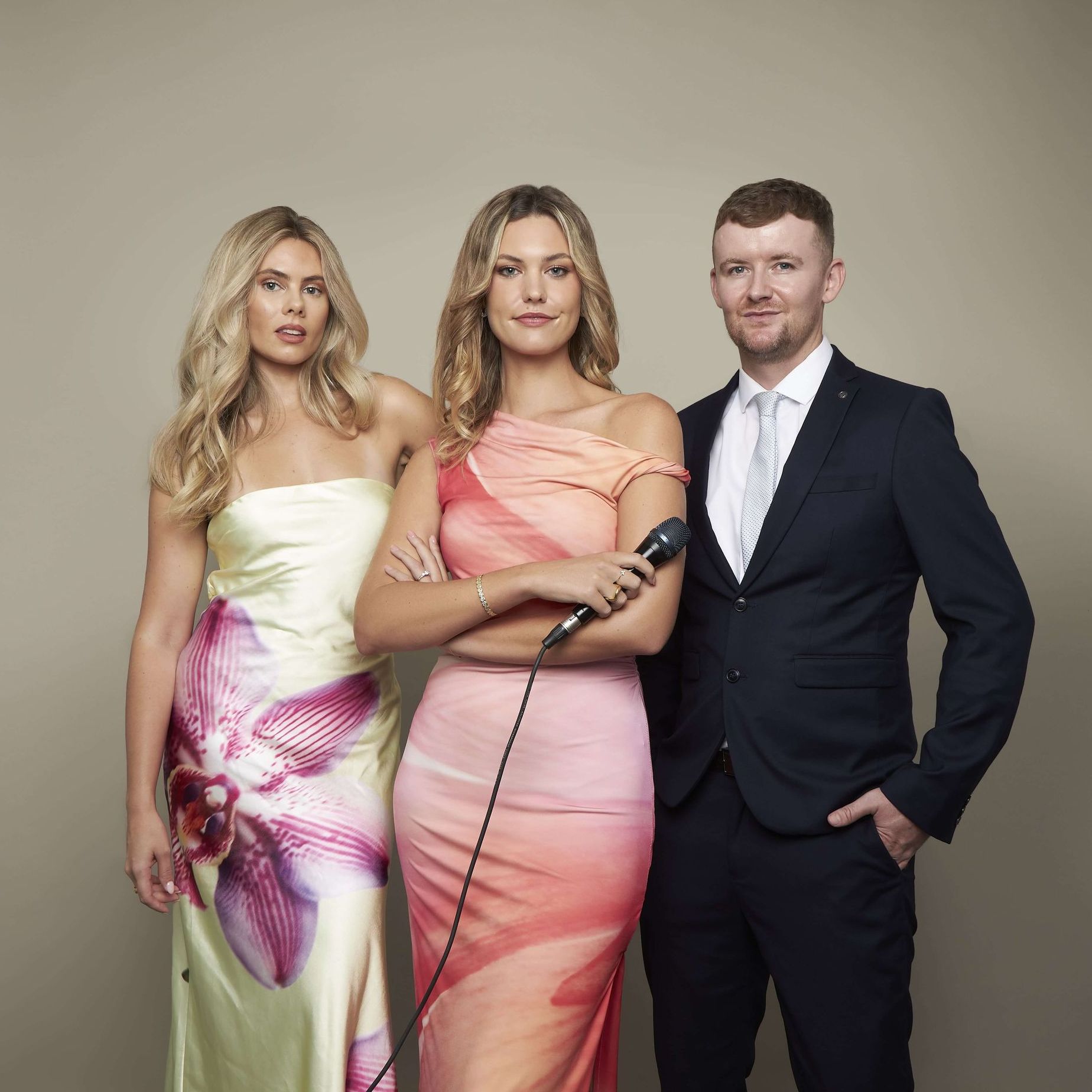 Two women in formal dresses and a man in a suit stand before a plain background; the center woman holds a microphone.