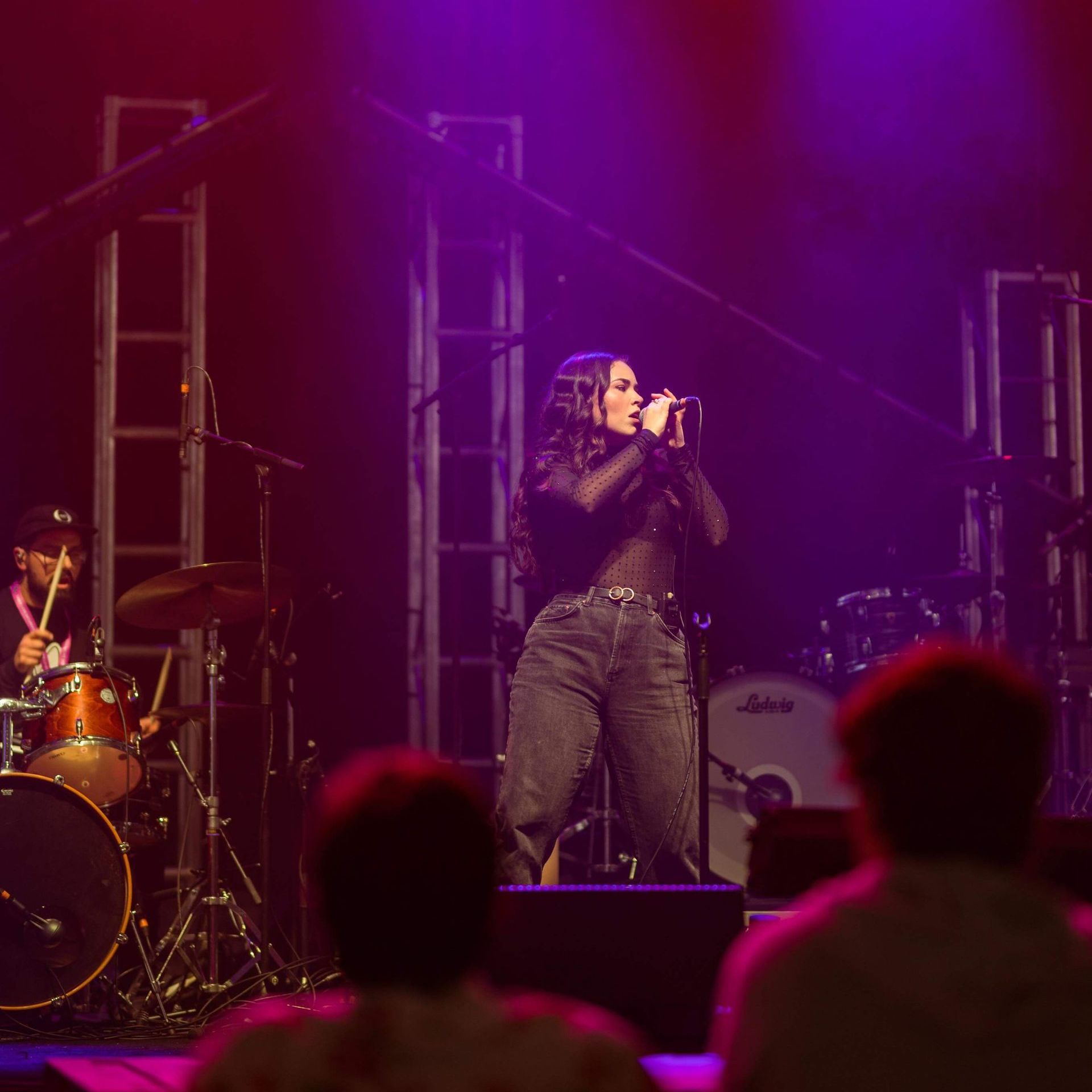 A singer performs on stage, holding a microphone, with a drummer and audience visible.