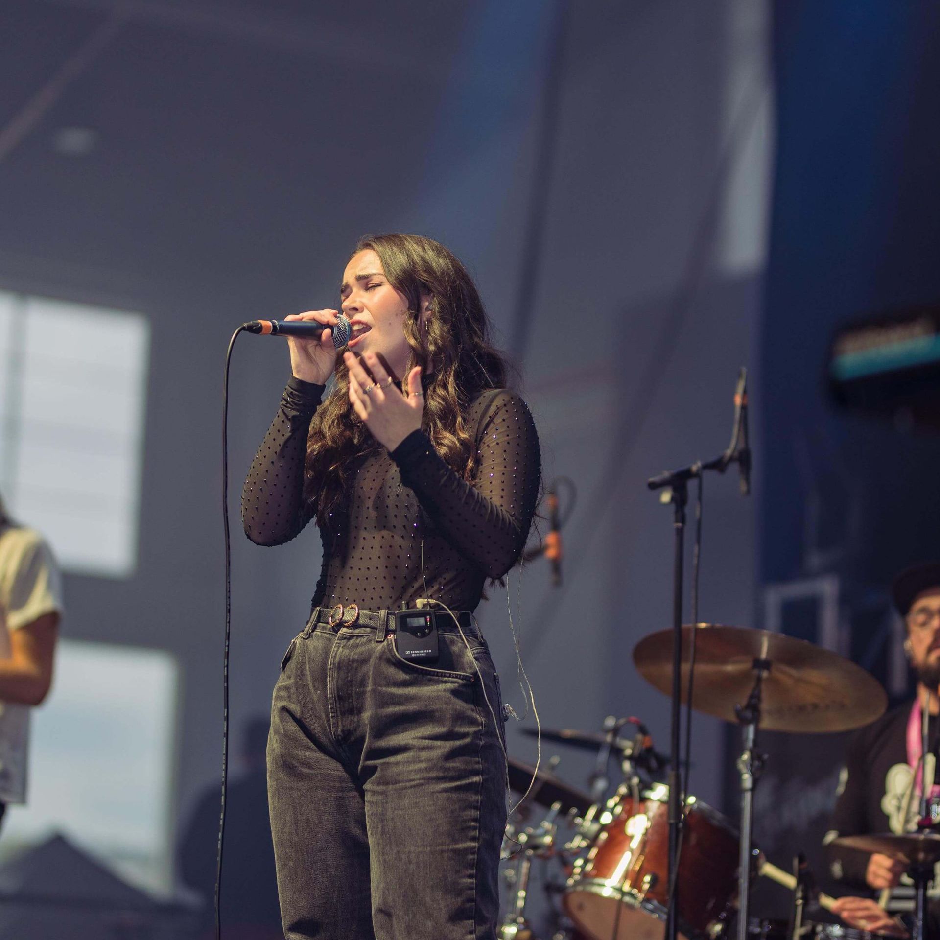 Woman singing into a microphone on stage, wearing a black top and jeans. Drummer visible in background.