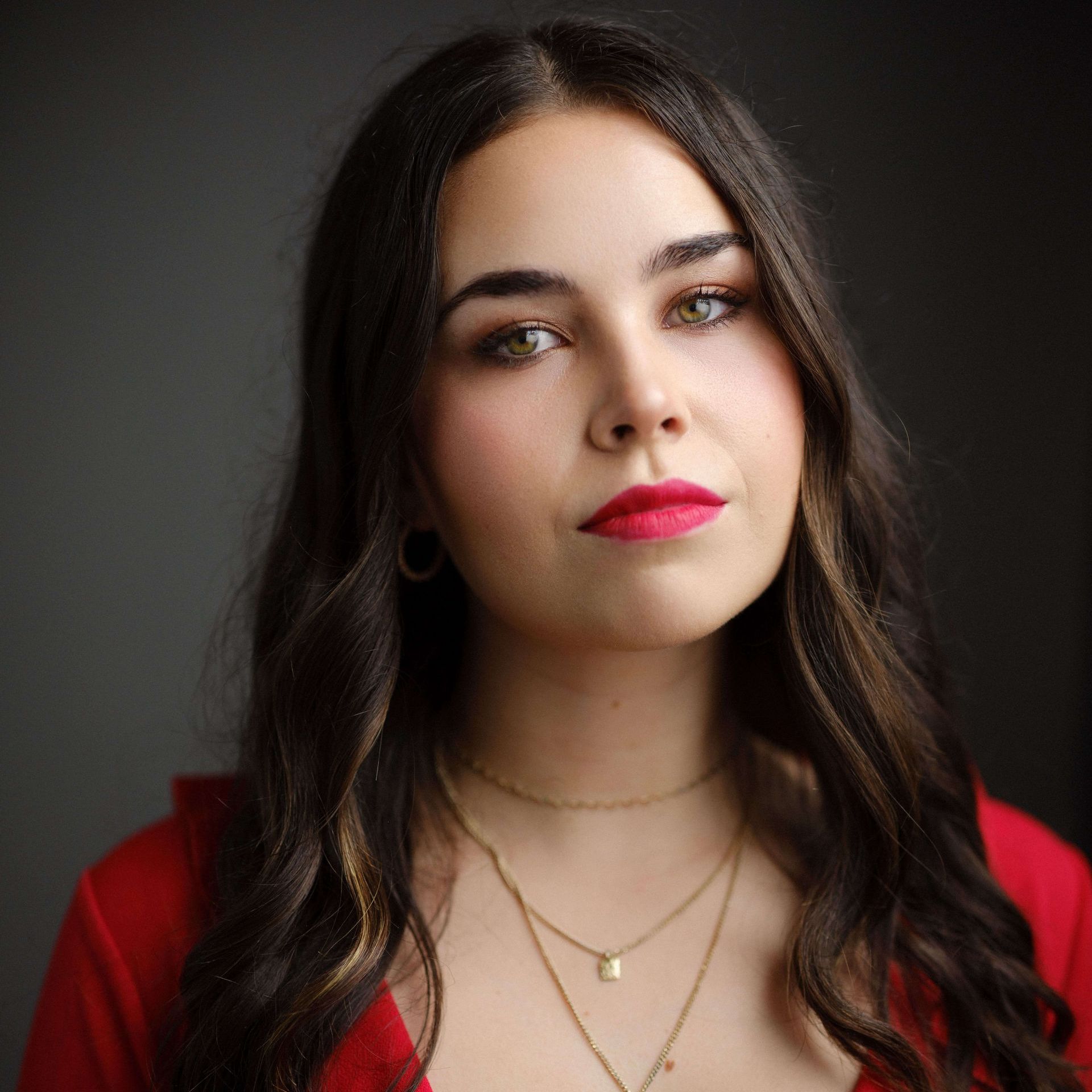 Woman with brown hair and red lipstick, wearing a red top and gold necklaces, looking forward.