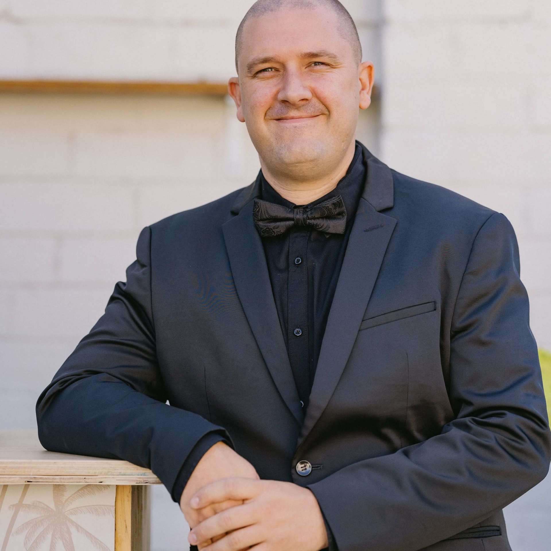 Man in a dark suit and bow tie leaning on a stone railing, smiling outdoors.