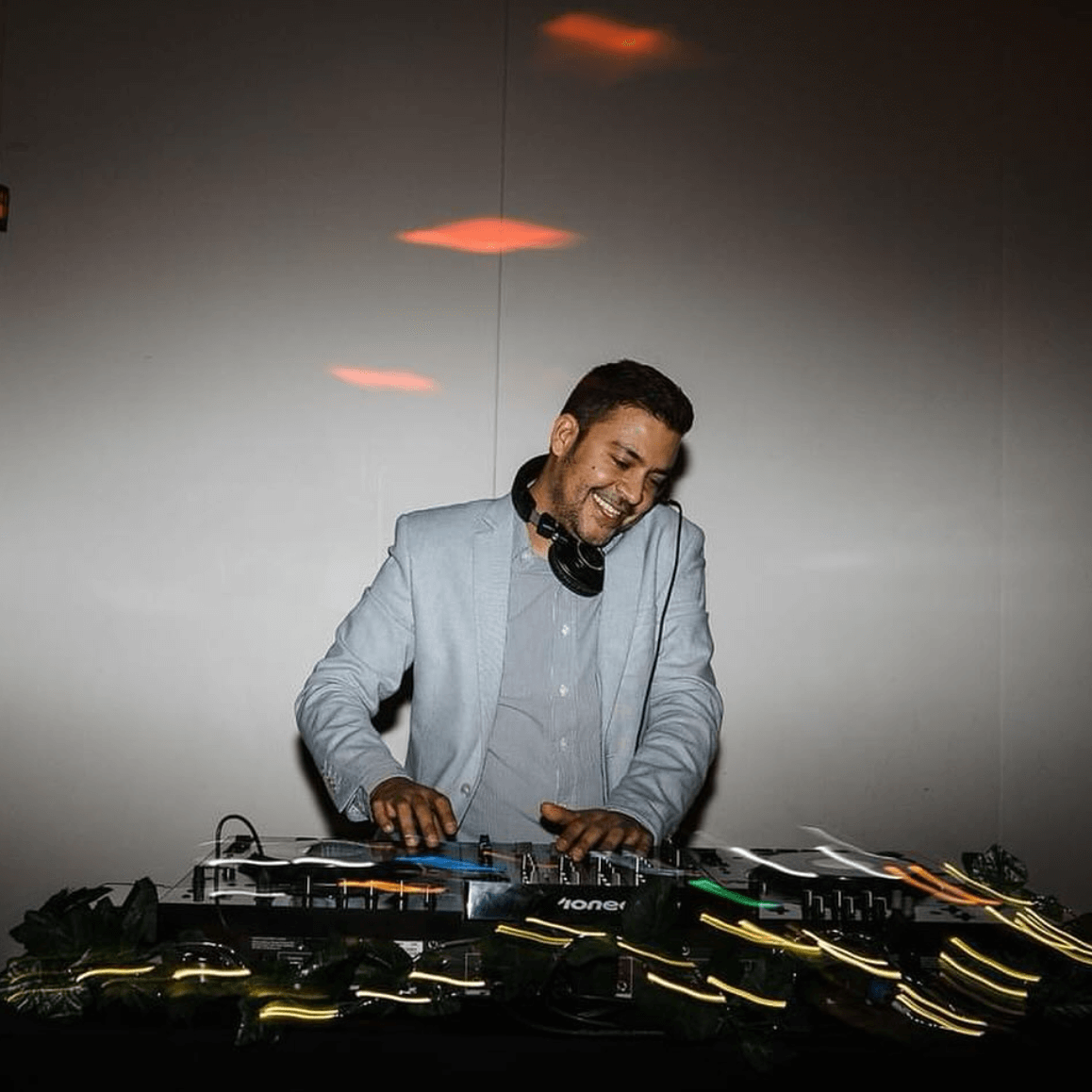 DJ wearing a light blue blazer and headphones, mixing music on a console, smiling.