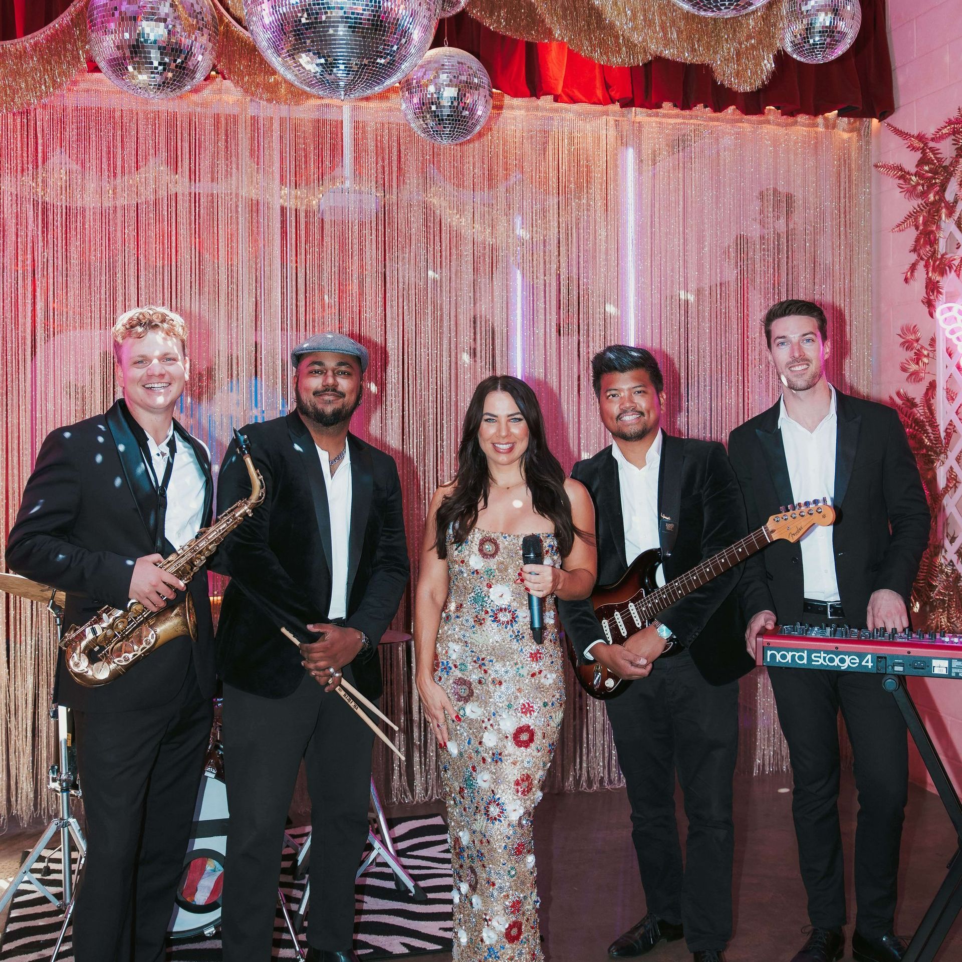 A five-piece band standing on stage with disco balls and gold fringe curtains, performing in formal attire.