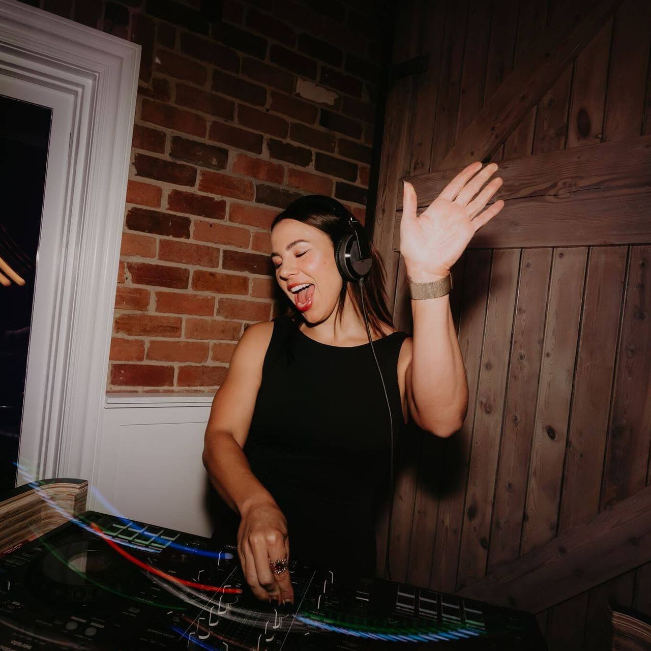 Woman DJ with headphones waves at a party, standing in front of mixing equipment near a brick wall.
