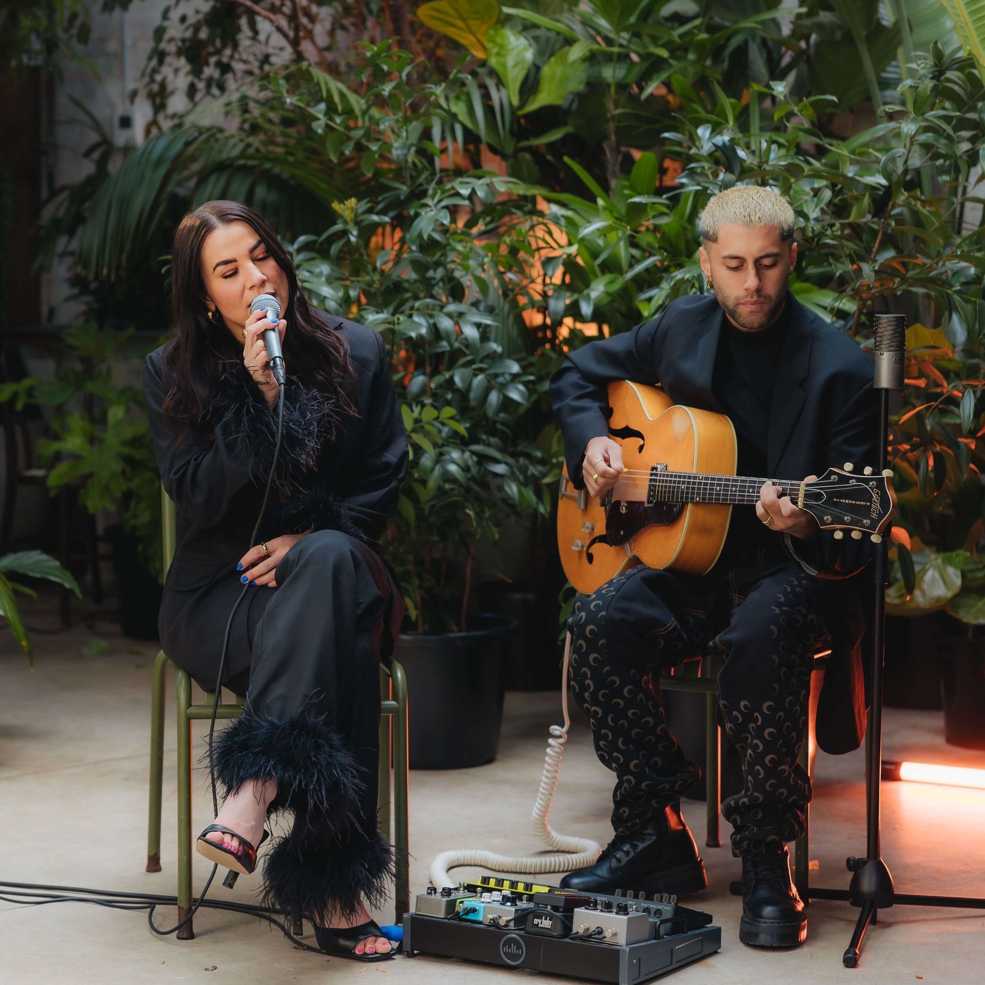 A singer and a guitarist perform in an indoor garden setting, seated on chairs with a pedalboard on the floor.