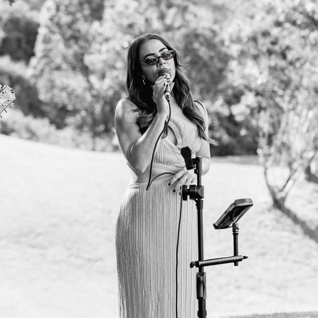 A person wearing sunglasses and a textured gown sings into a microphone on a stand in a sunlit, outdoor setting.