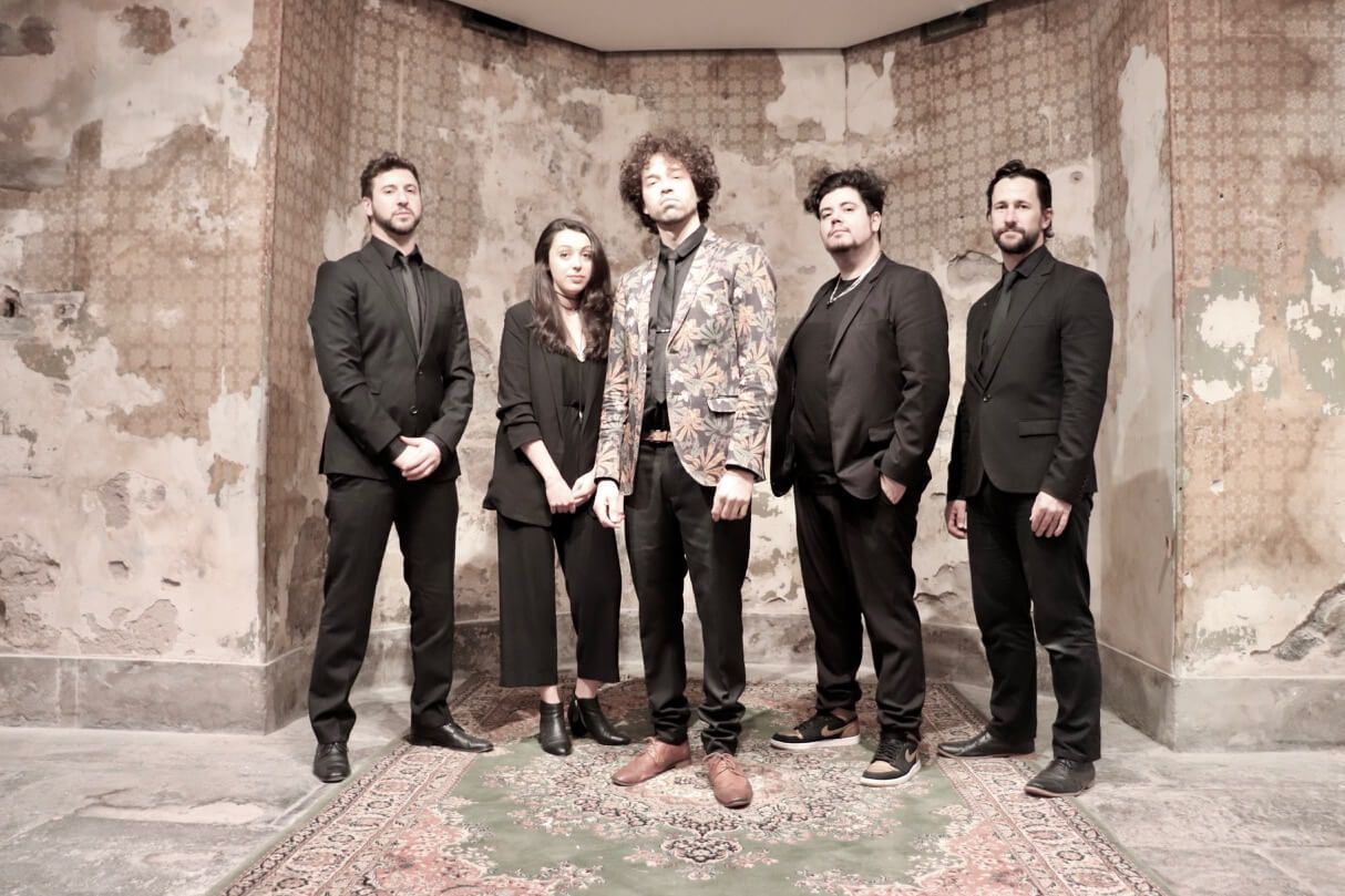 A musical band stands in a room. The lead singer is in a patterned jacket, others in black. They pose against a textured wall.