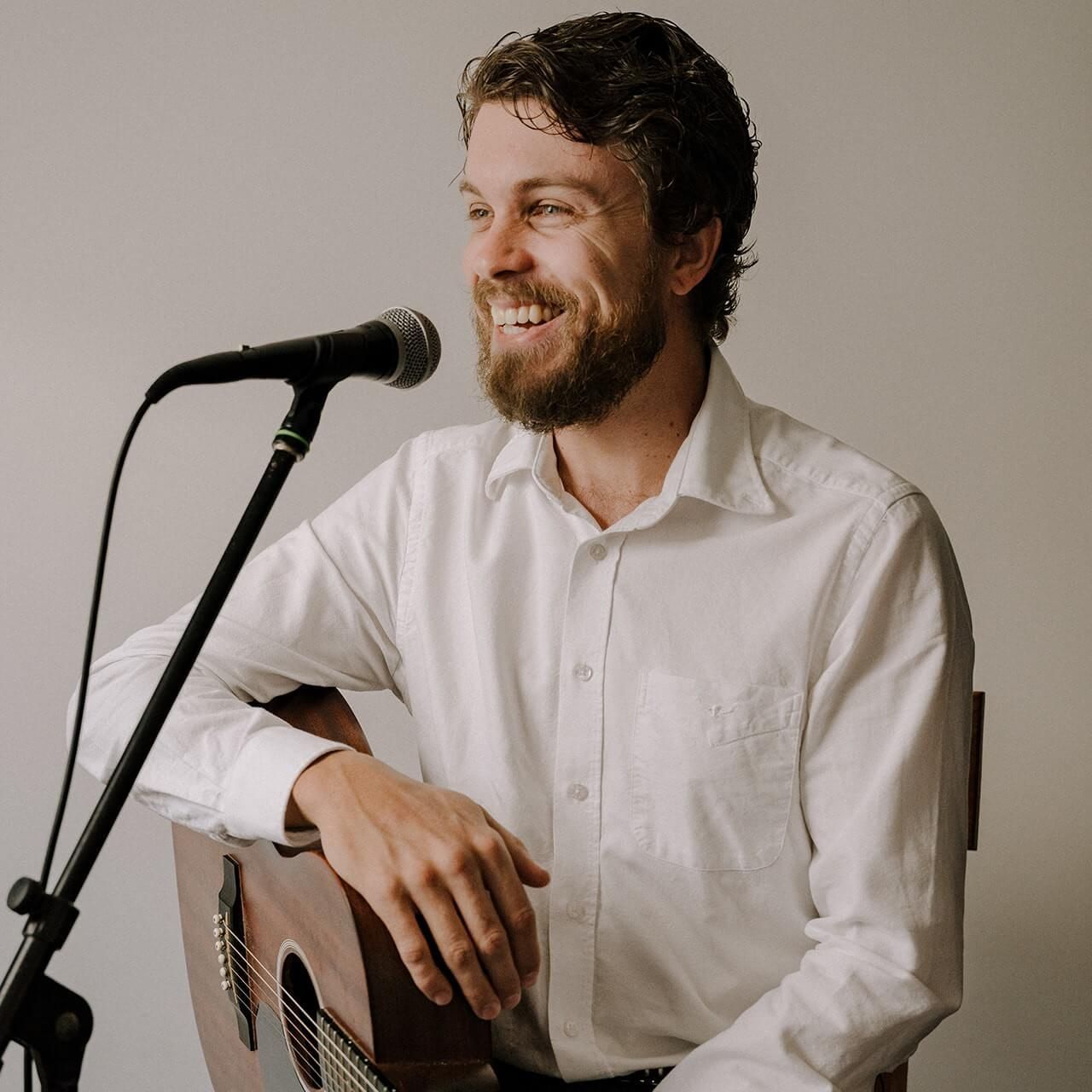 A person with a beard, wearing a white button-down shirt, smiling while holding an acoustic guitar near a microphone.